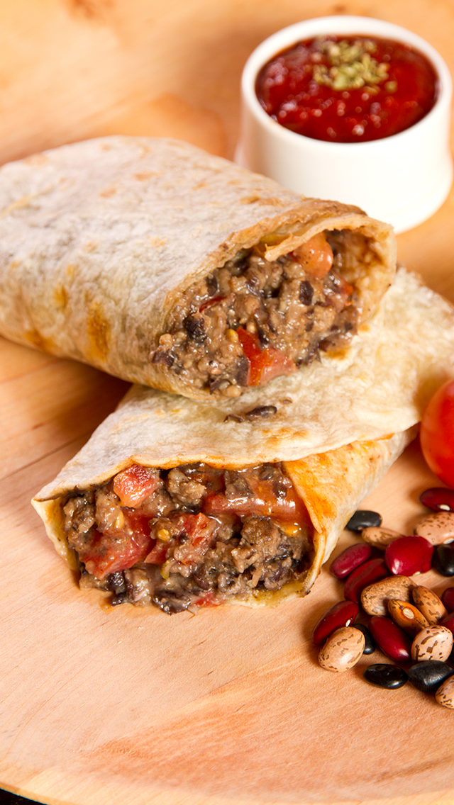 Two beef and bean burritos on a wooden cutting board, one cut open to reveal meat, beans, and vegetables, with a side of tomato soup garnished with herbs and some scattered beans.