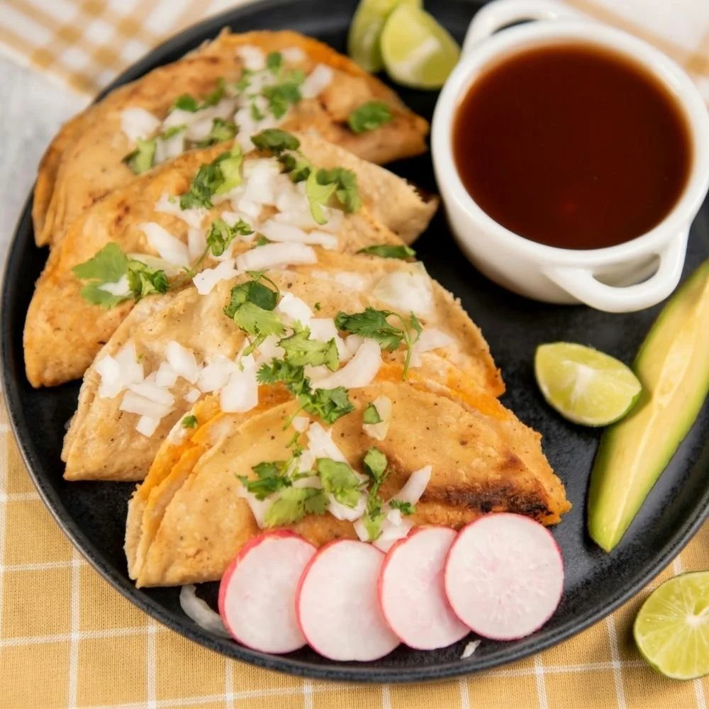 A plate of three crispy tacos topped with chopped onions and cilantro, served with lime wedges, sliced radishes, and a cup of dark sauce.