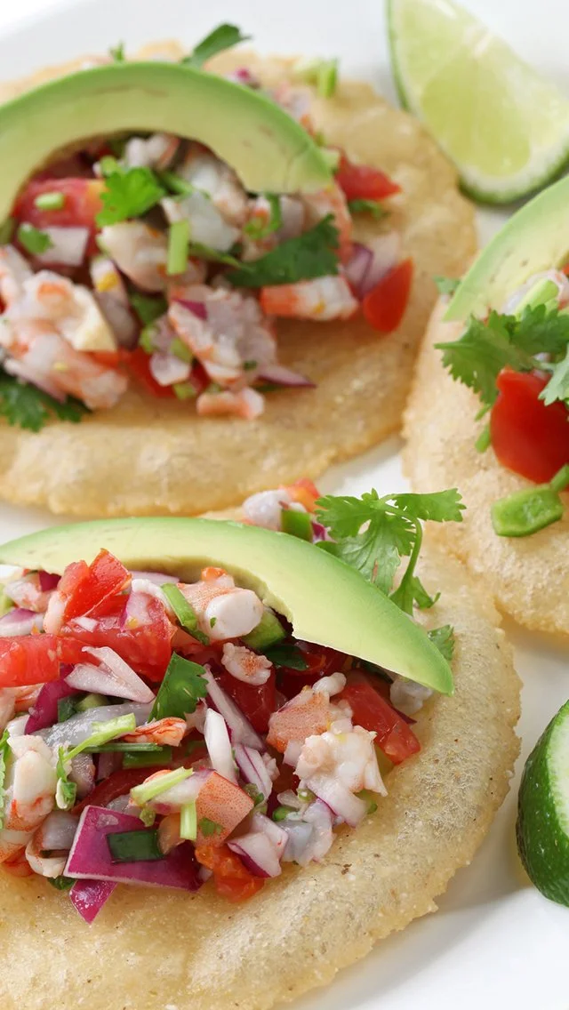 Three crispy fish tacos topped with diced tomatoes, chopped onions, cilantro, and sliced avocado, served with lime wedges.