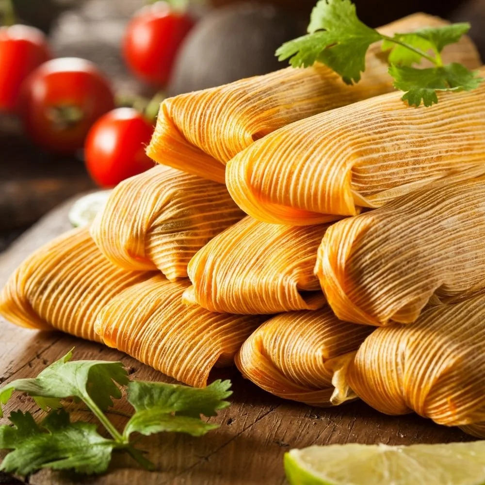 Stack of tamales garnished with cilantro, with tomatoes and lime in the background.