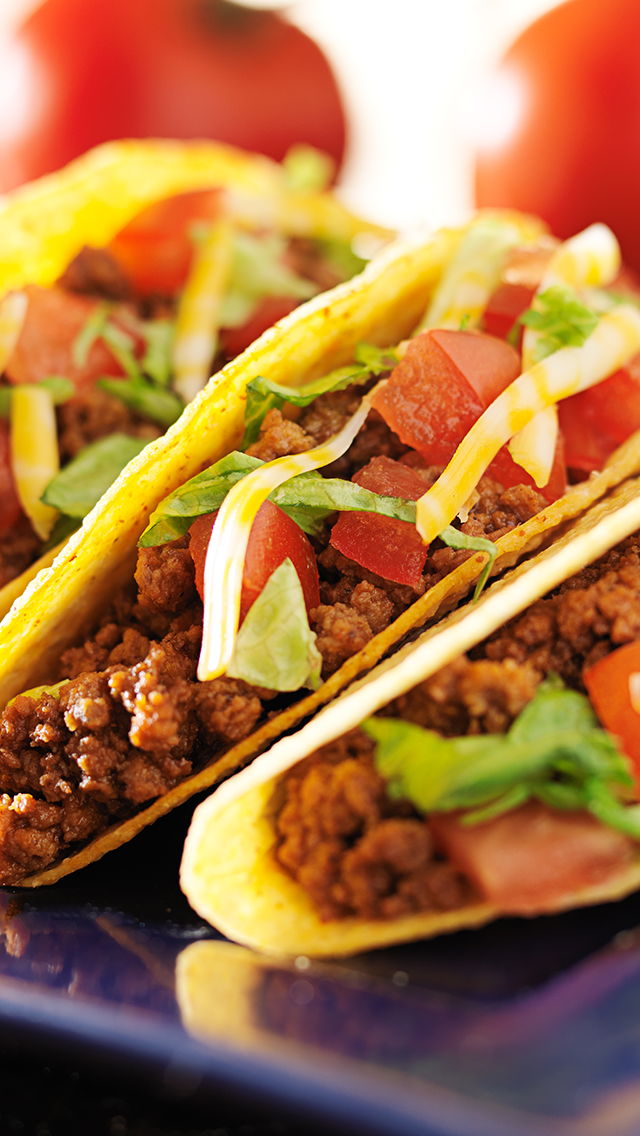 Three tacos filled with seasoned ground beef, chopped tomatoes, shredded cheese, chopped lettuce, all in corn taco shells, on a black plate.