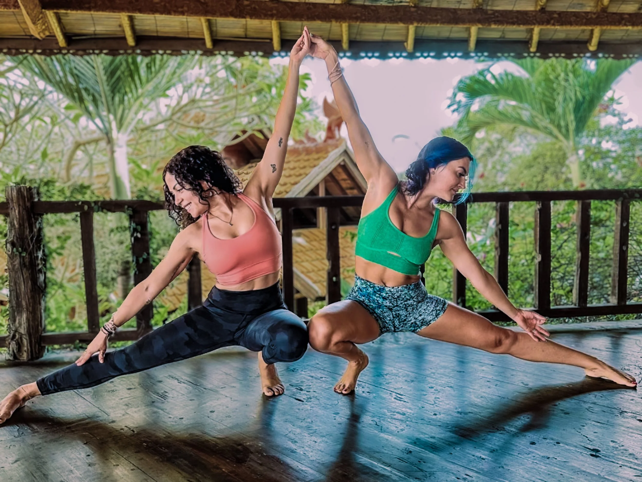 Ashlee and Kimmy in a mirrored yoga lunge with hands joined overhead on an open-air jungle deck
