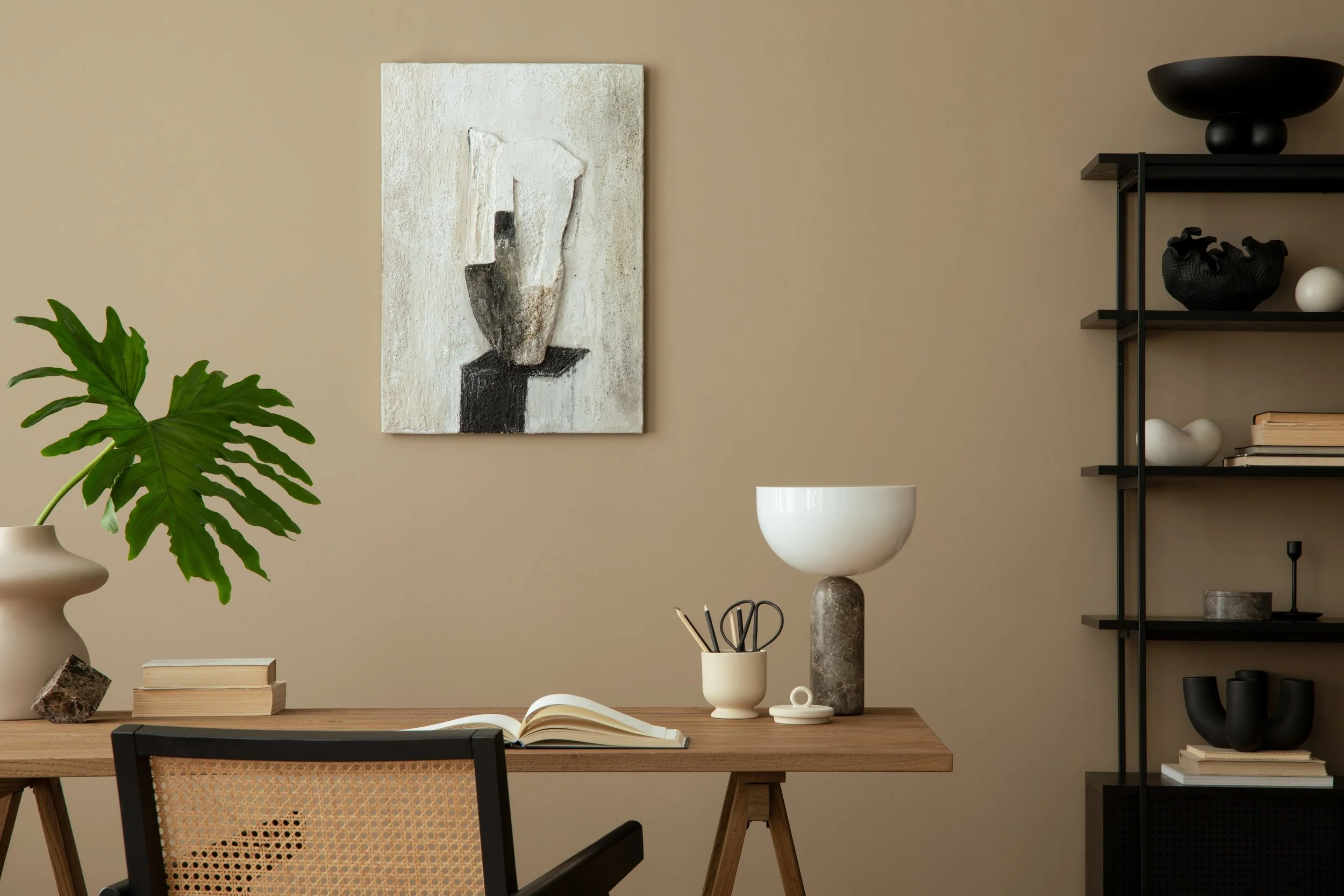 Minimalist home office with a wooden desk, open book, ceramic bowl filled with pens and scissors, small notepad, and large decorative lamp. A green leafy plant in a beige vase is on the left, abstract art hangs on the beige wall, and black shelves with decorative objects are on the right.