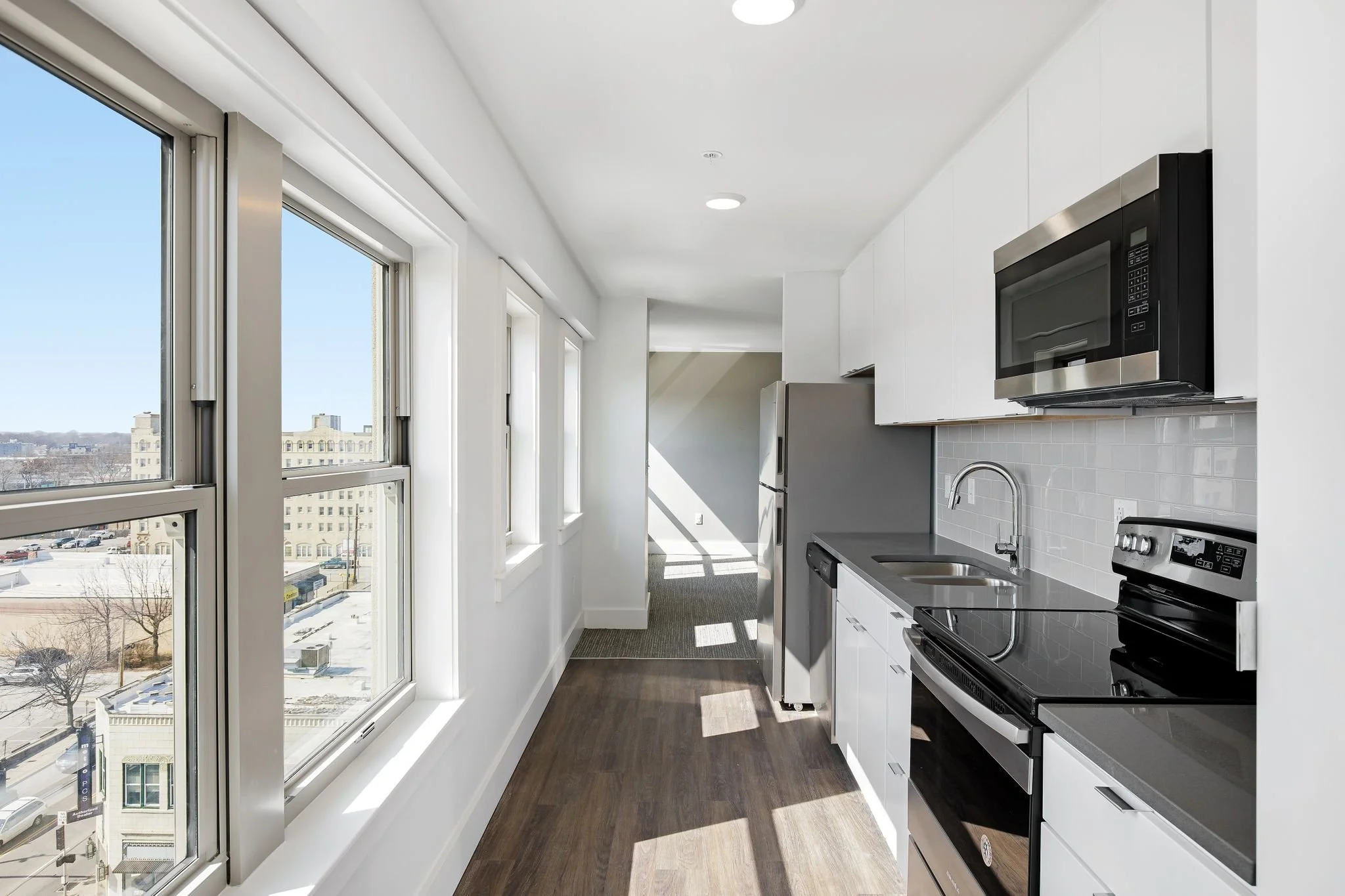 Modern kitchen with white cabinets, gray countertops, stainless steel appliances, a window with a city view, and wood flooring.