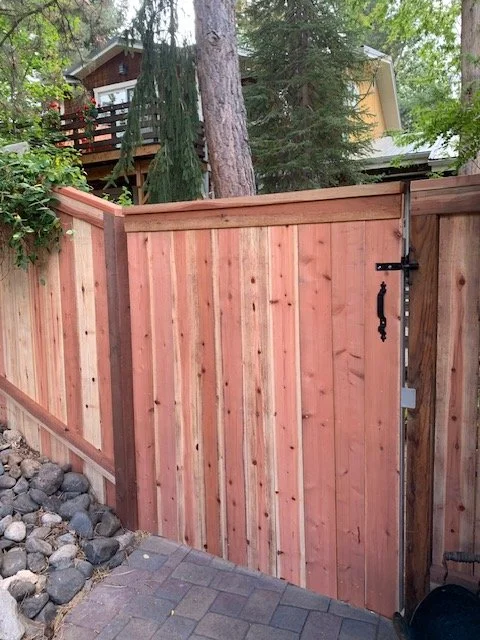 Wooden fence with a gate, set in a backyard with trees and a house in the background.