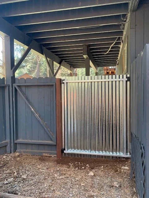 A fenced area with a metal gate, under a wooden deck with exposed beams.
