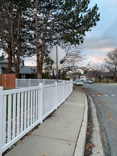 Empty sidewalk next to a white fence on a residential street with parked cars and trees in the background.