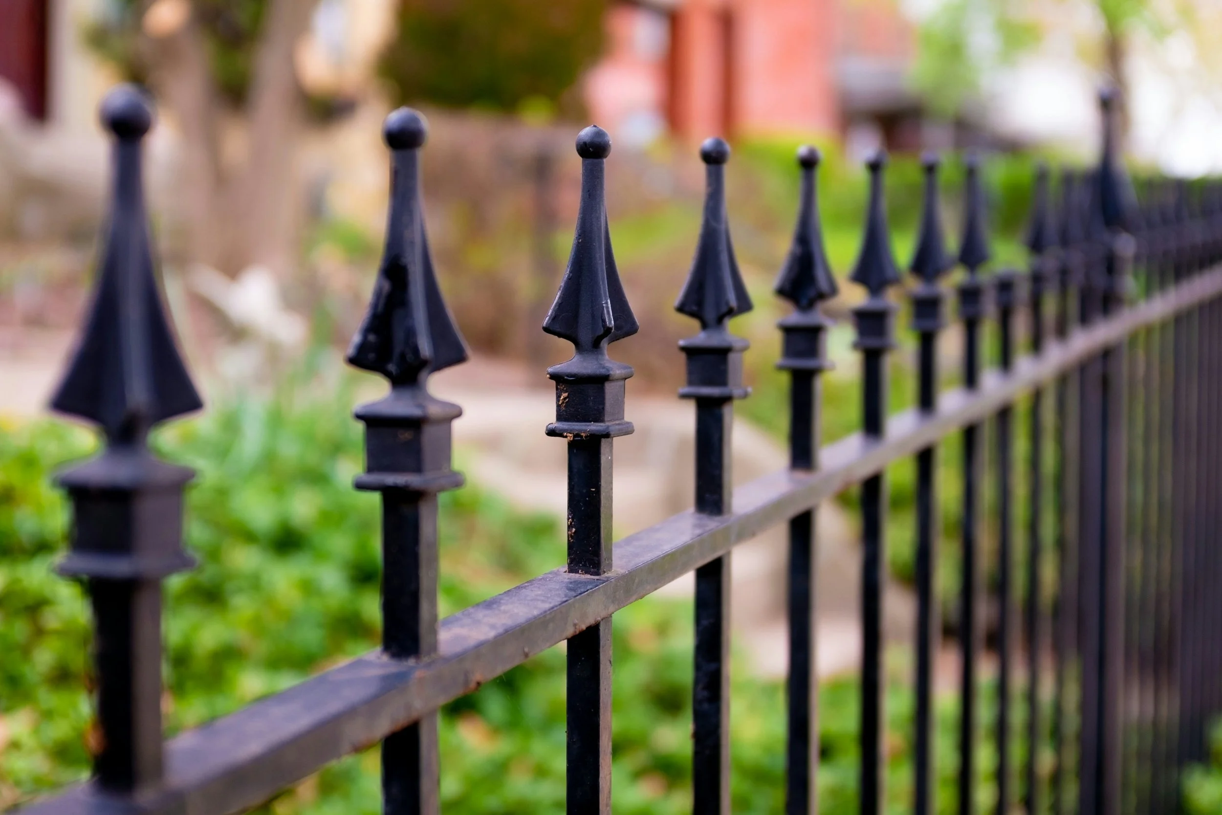 Close-up of a black wrought iron fence with pointed finials, with a blurred background of greenery and houses.