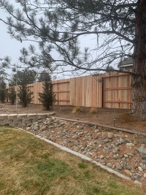 Backyard with a new wooden fence, a small stone retaining wall, and a dry creek bed with rocks and pebbles, trees, and a grassy lawn.