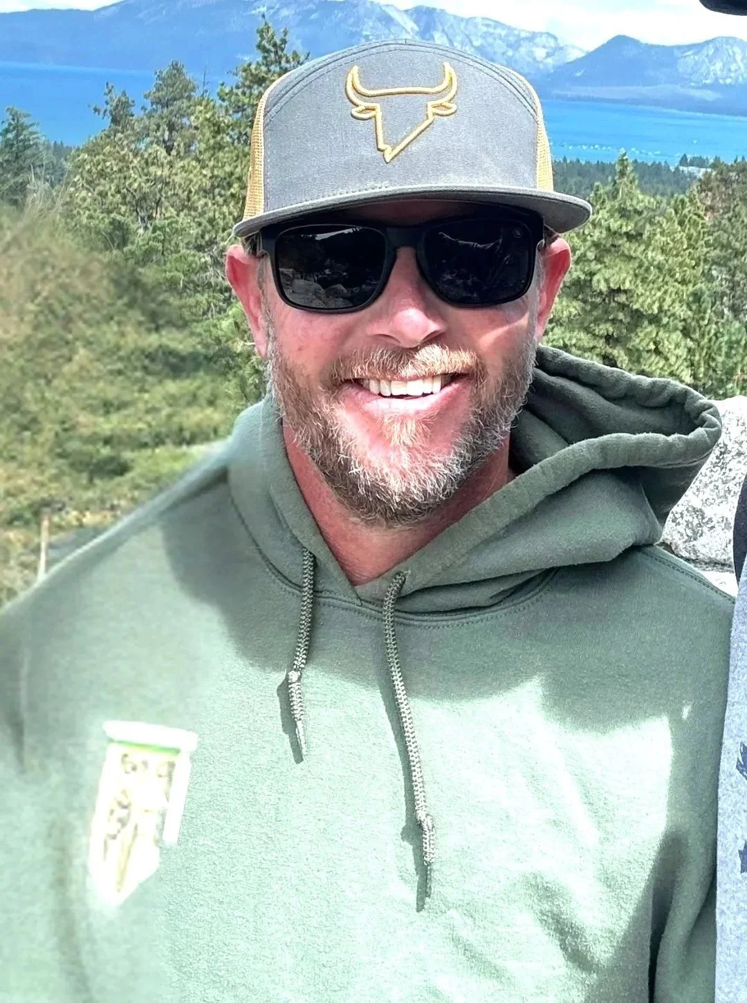 A man with a beard and sunglasses smiling outdoors, wearing a gray baseball cap with a logo, a green hoodie, and in the background, a scenic view of trees, mountains, and a lake.
