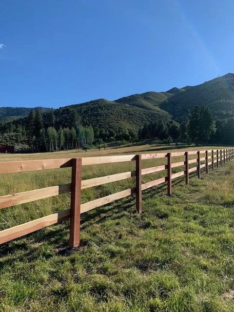 A wooden fence running through a grassy field with mountains and a clear blue sky in the background.