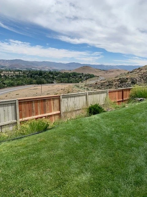 A backyard with green grass and a wooden fence overlooking a hilly landscape with mountains and a partly cloudy sky.