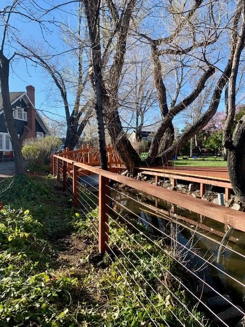 A wooden fence runs along a waterway next to a walking path, with leafless trees and some houses in the background on a clear day.