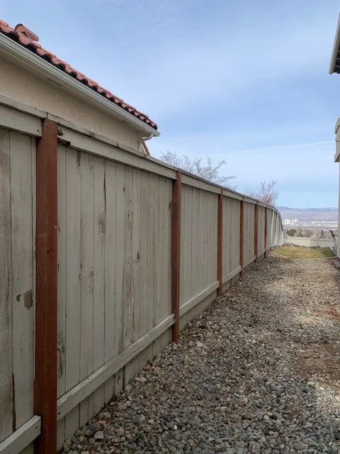 A long wooden fence runs along the side of a house with a red tile roof, with a gravel path beside it. The sky is cloudy, and there are trees and a distant landscape in the background.
