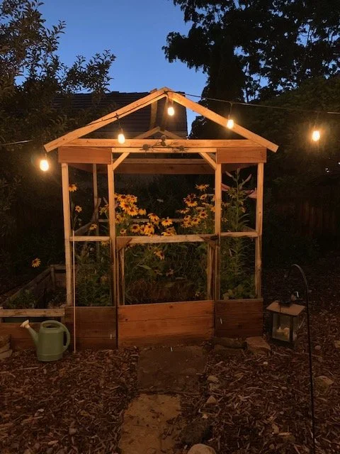 A small wooden garden shed with string lights hanging from the roof at dusk, surrounded by flowers and trees.
