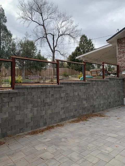 A backyard with a brick wall, a stone patio, and a new glass railing on top of the wall, with trees and house in the background.