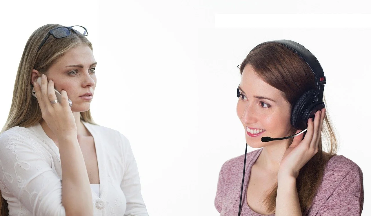 Two women talking on phones, one with a headset and the other holding her phone to her ear, against a plain background.