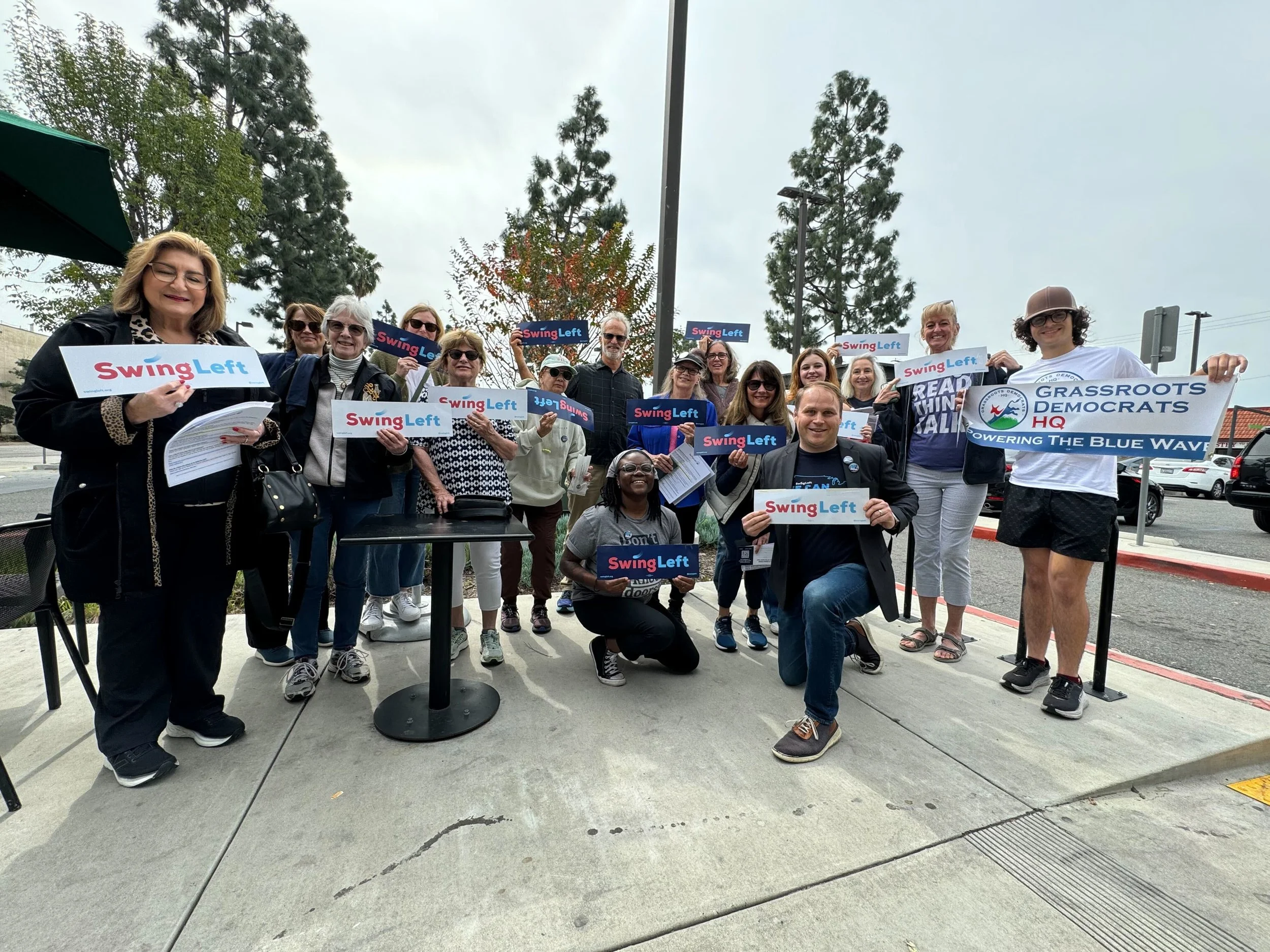 A diverse group of people gathered outdoors during the daytime, holding signs that read 'Swing Left' and a larger banner that says 'Grassroots Democrats HQ.' The group appears to be at a political or community rally, with trees and a parking lot in the background.