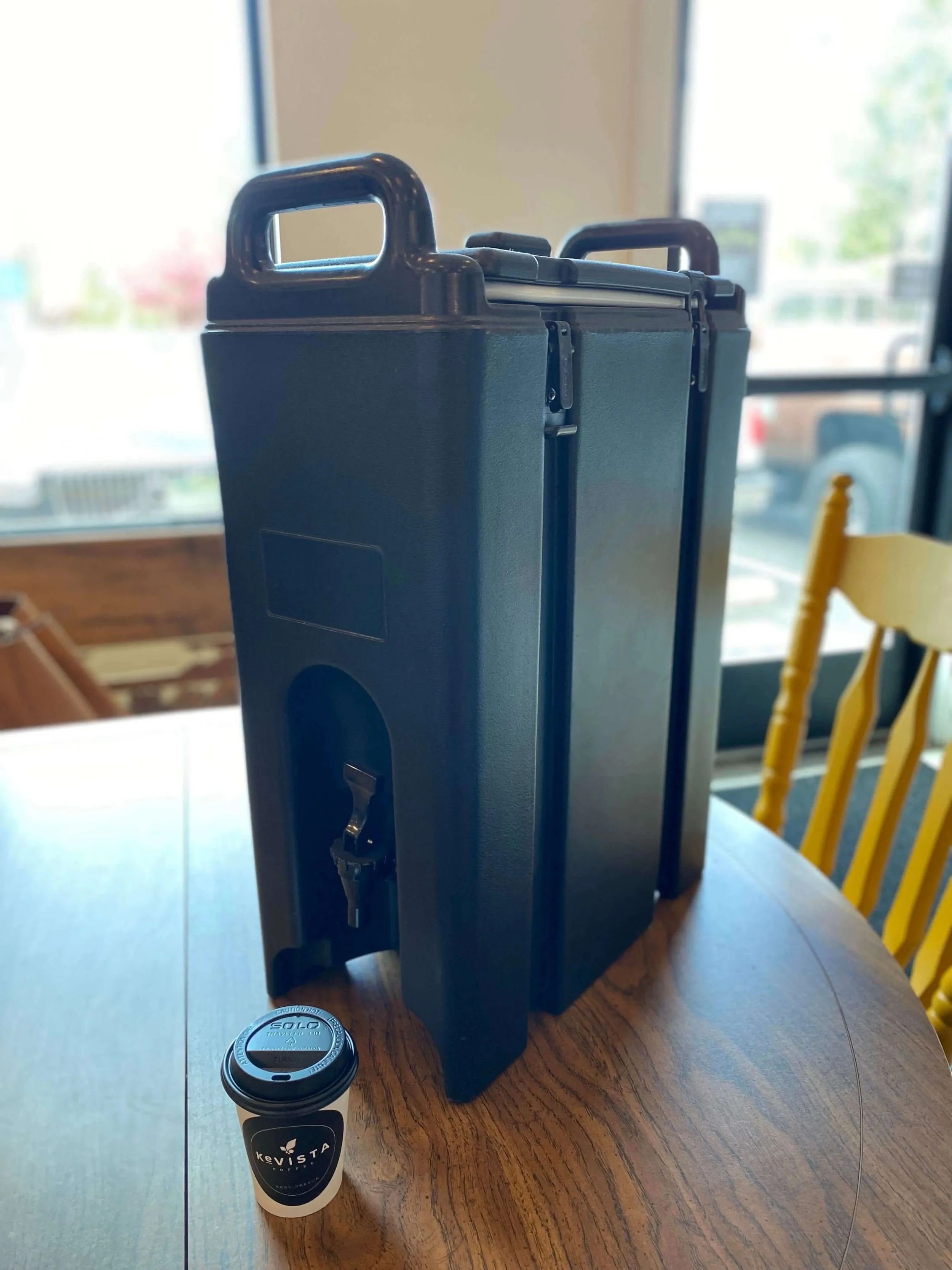 Large black beverage dispenser with spout on a table next to a small coffee cup.