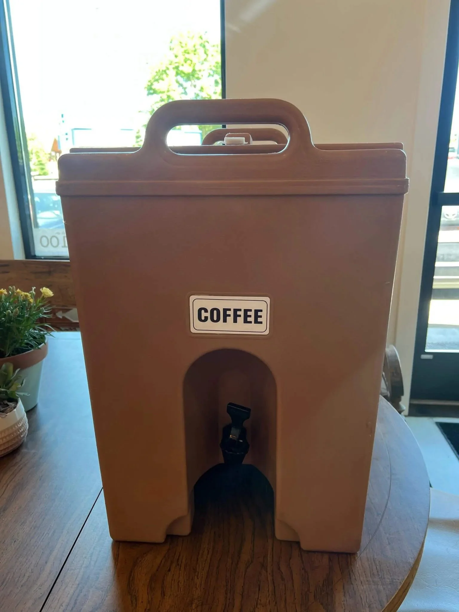 Brown insulated beverage dispenser labeled "Coffee" on a wooden table indoors.
