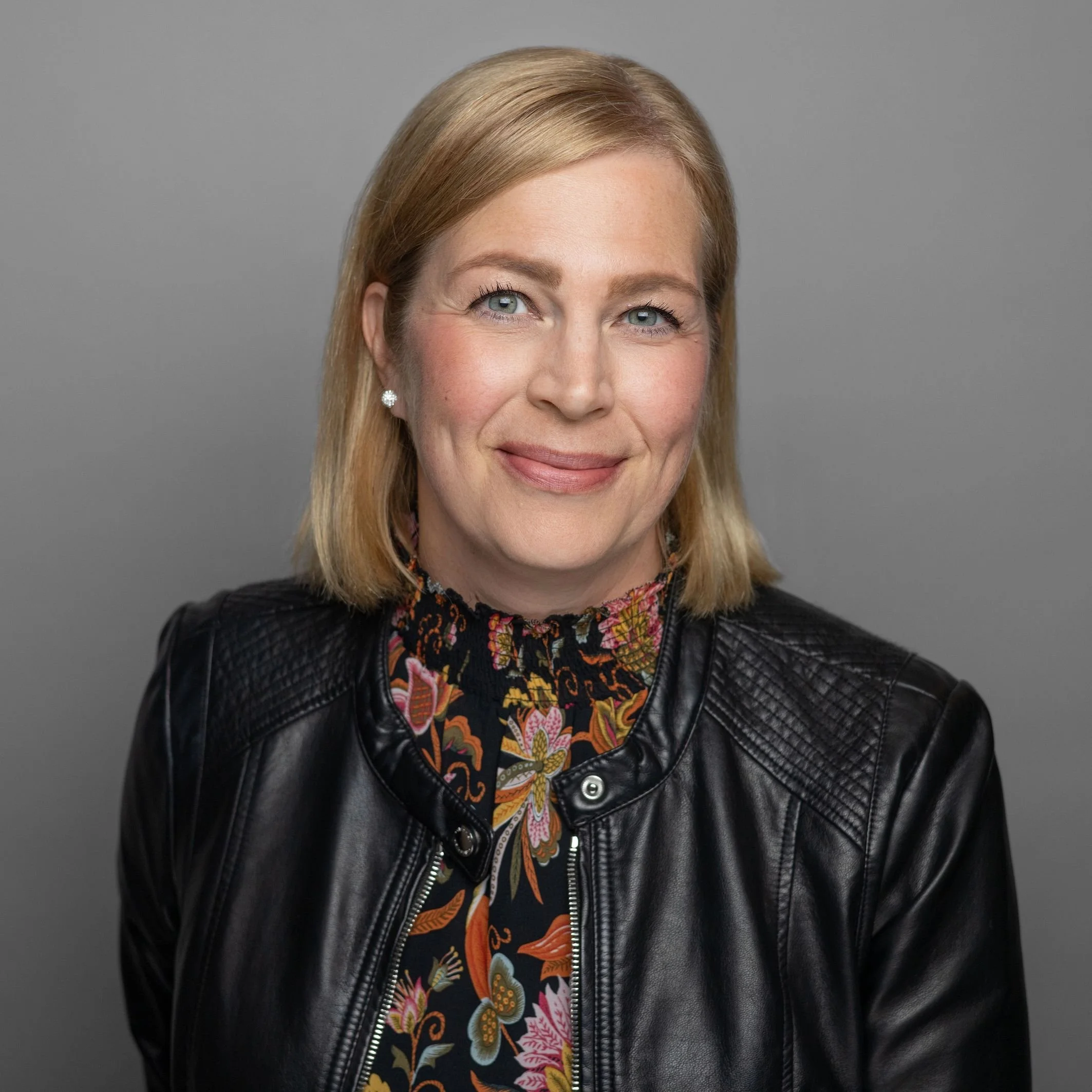 A close-up of a woman's face with blonde hair, blue eyes, red lipstick, and earrings, wearing a black jacket and a colorful patterned shirt against a gray background.