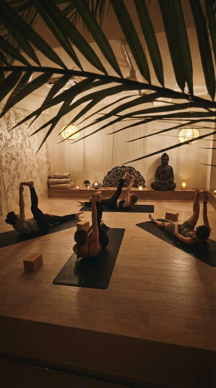People practicing yoga in a peaceful, dimly lit studio with candles and Buddha statue, covered partially by palm leaves in the foreground.