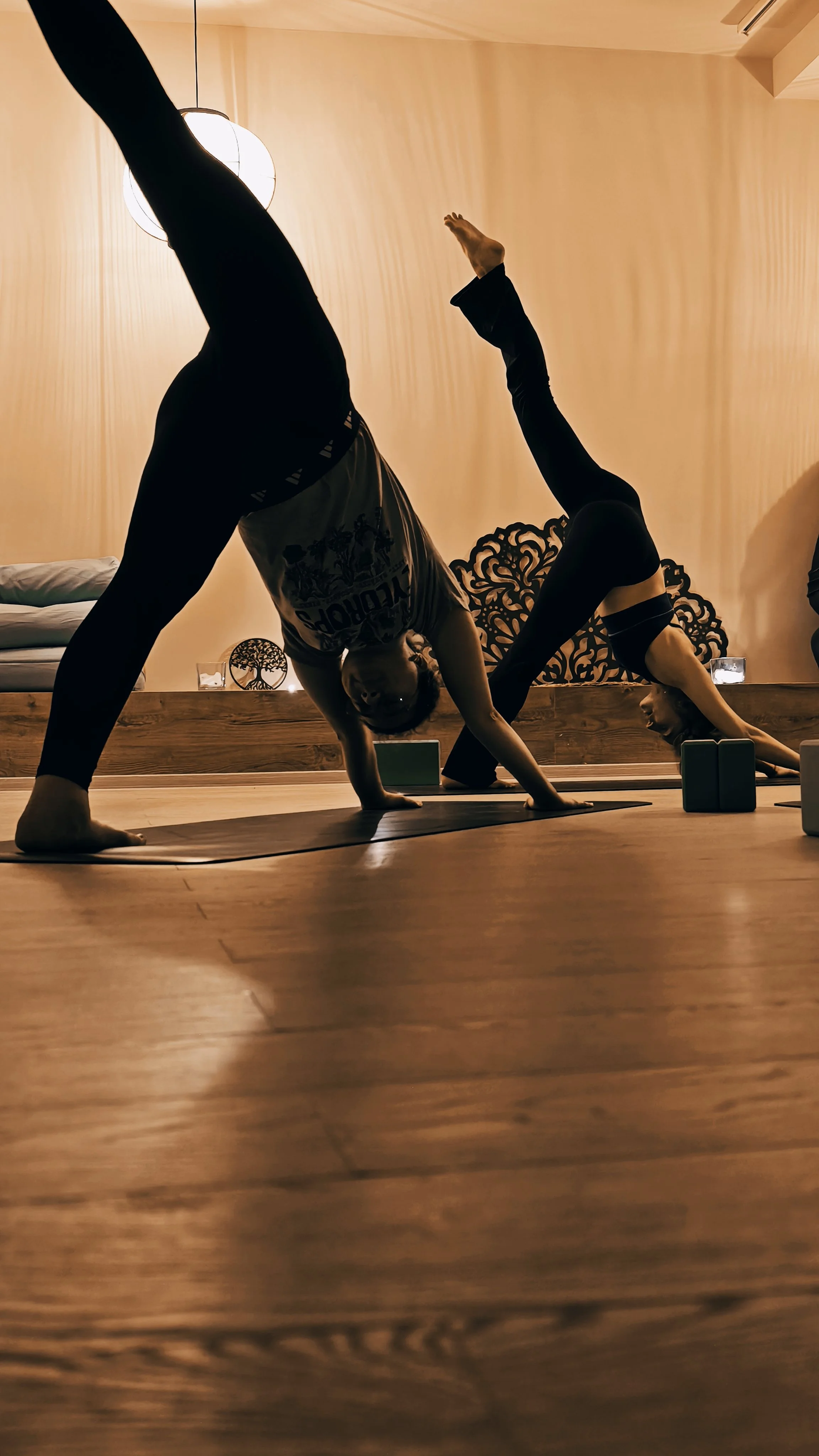 Two people practicing yoga in a dimly lit room, one in a downward dog pose and the other in a bridge pose, with decorative wall art and candles in the background.