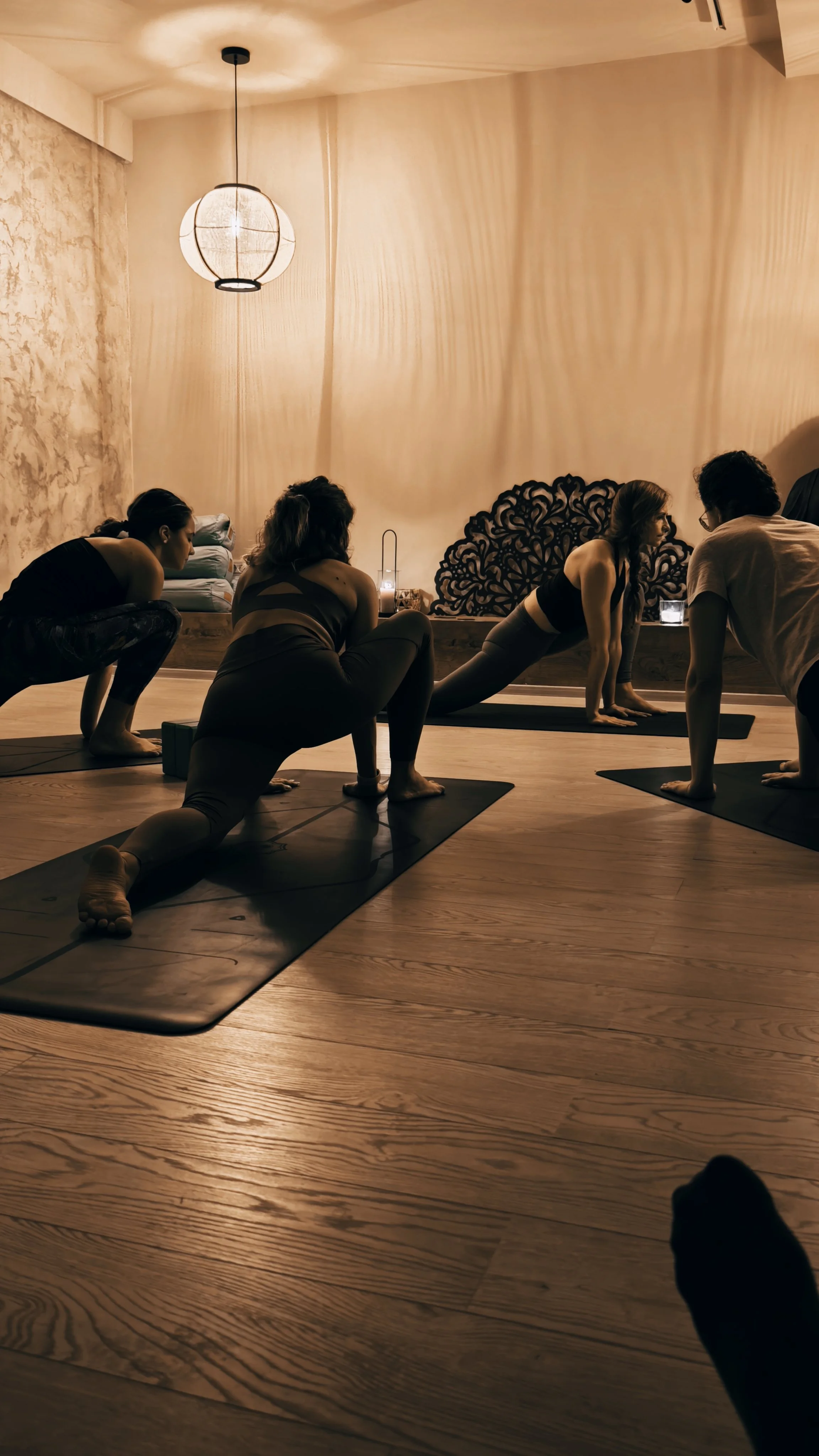 A group of people in a yoga class practicing yoga poses on mats in a dimly lit room with wooden floors and soft lighting, including a hanging paper lantern