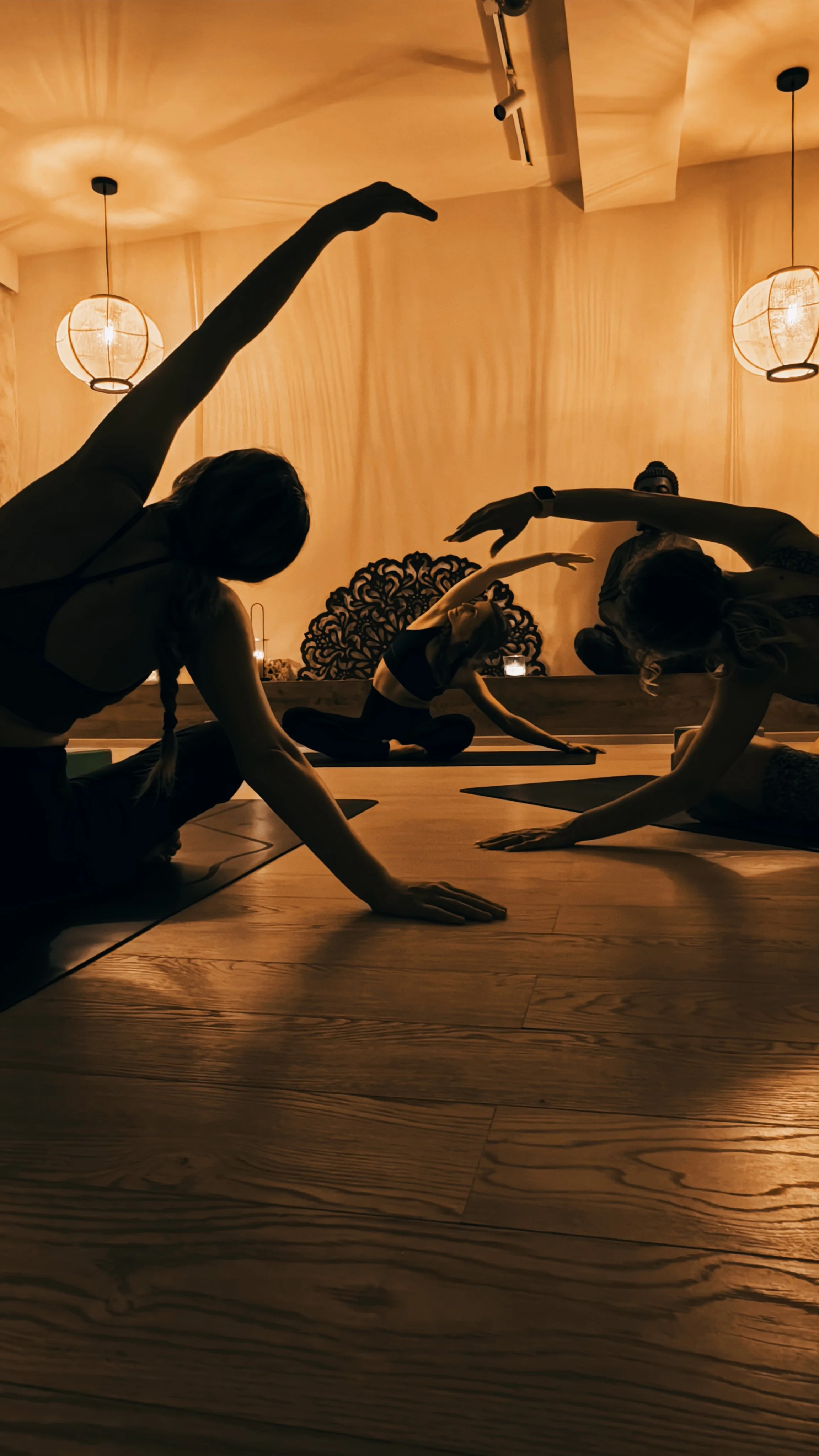 People practicing yoga in a dimly lit studio, seated on yoga mats with one arm extended overhead and the other hand on the floor, in a grappling pose.