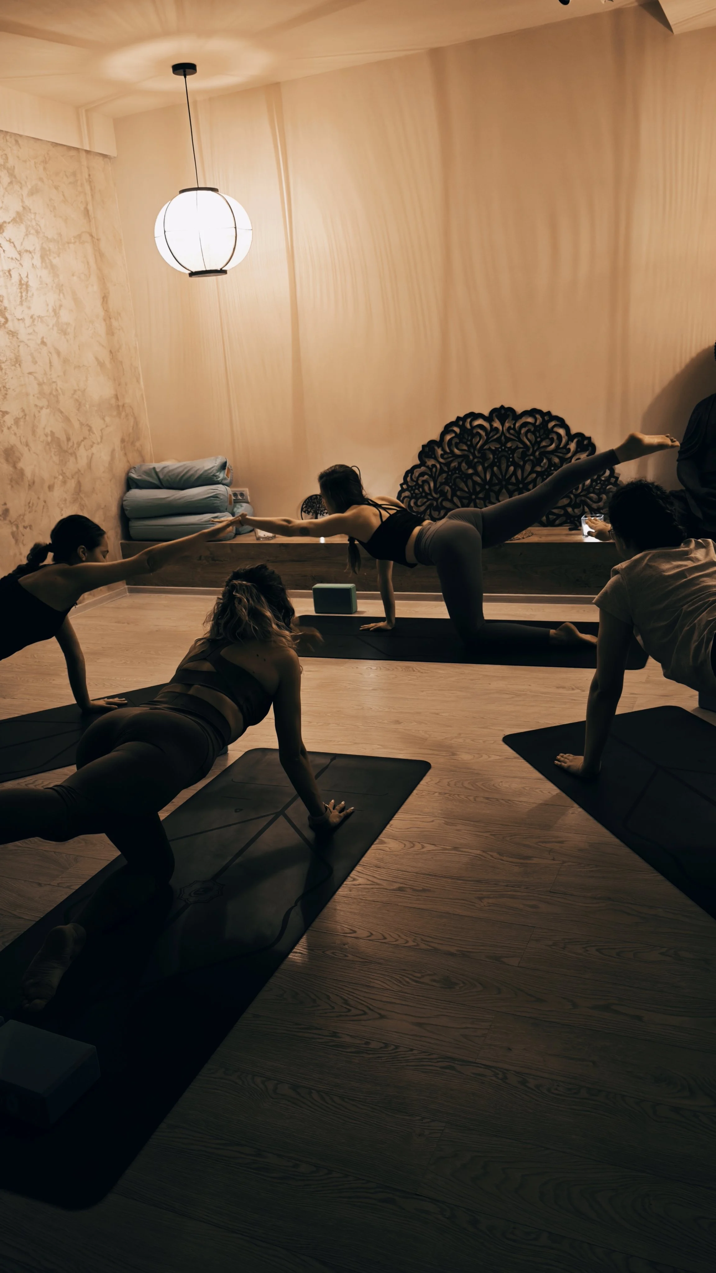 Women practicing yoga in a studio, performing a balance pose on yoga mats with dim, warm lighting.