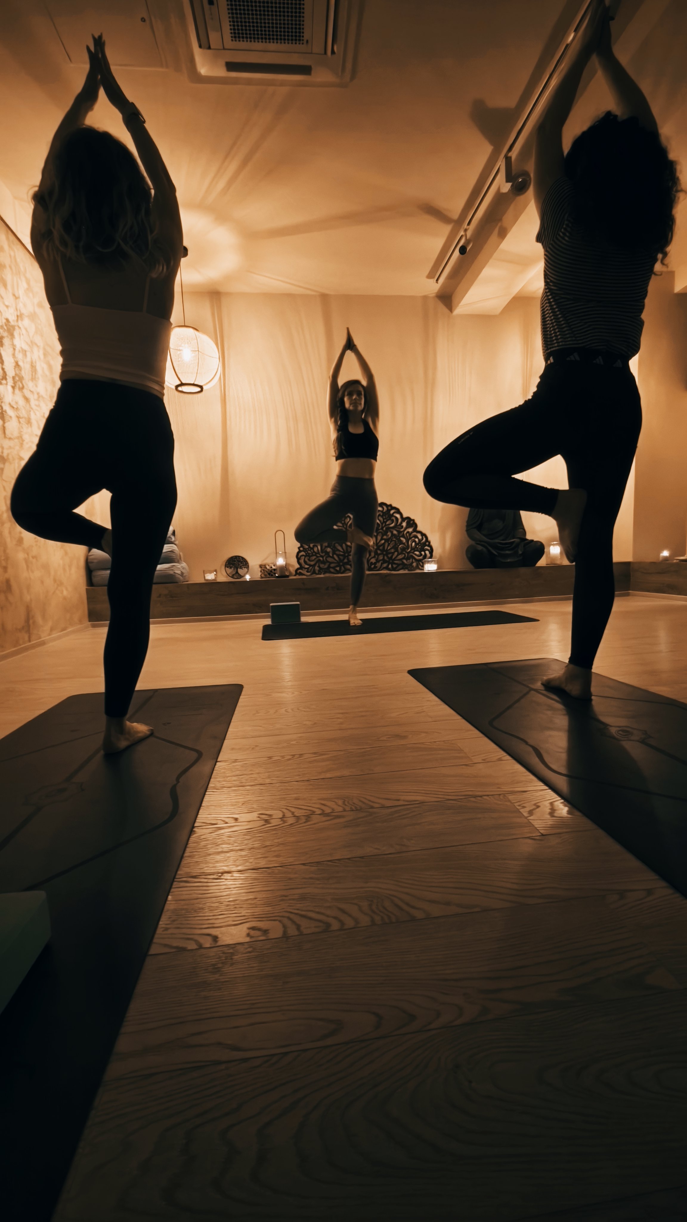 Three women practicing yoga in a dimly lit room, performing tree pose on yoga mats.