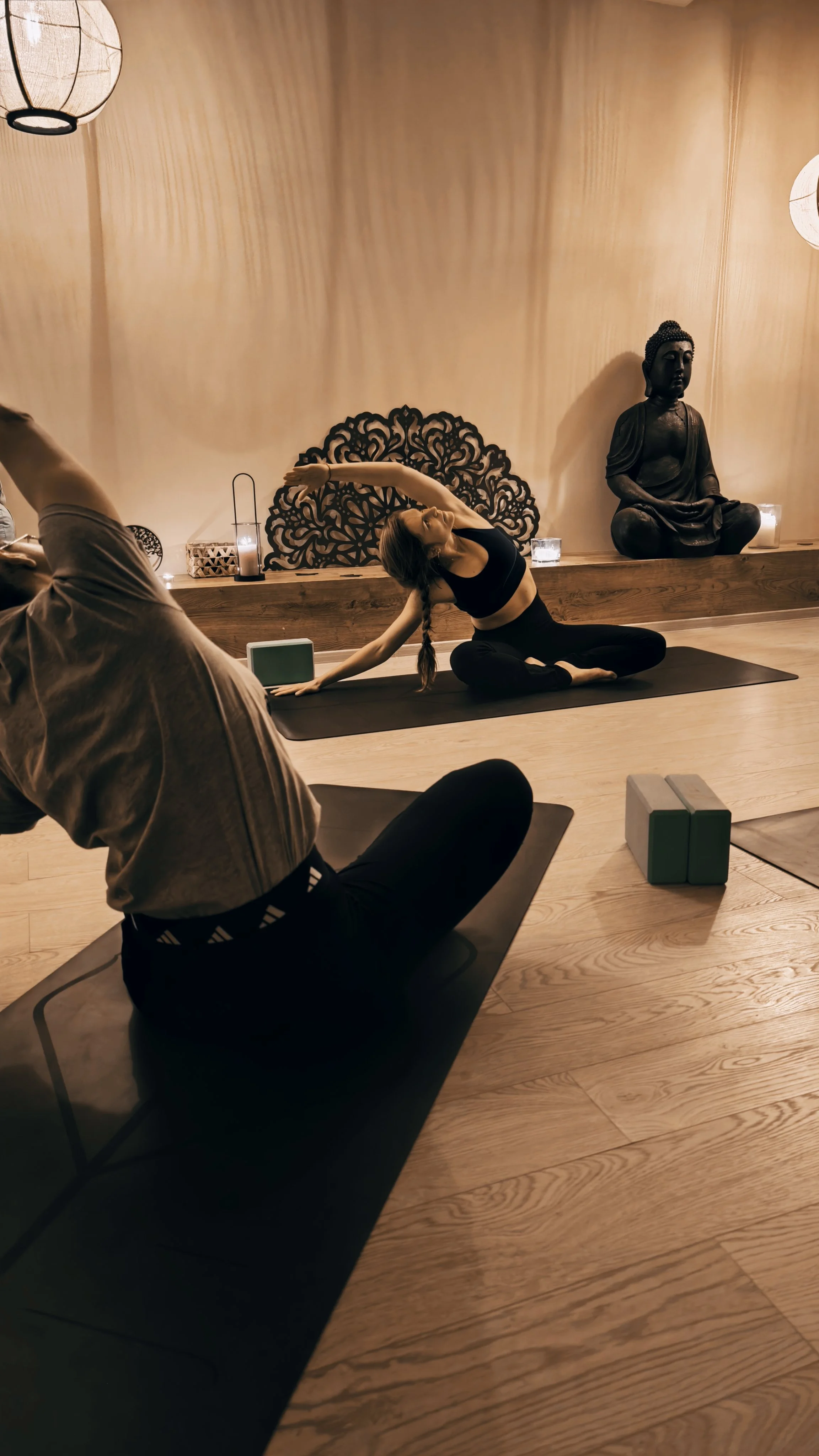 A woman practicing yoga in a dimly lit room with wooden walls, sitting on a black yoga mat, performing a side stretch. Buddha statues and candles are visible in the background.