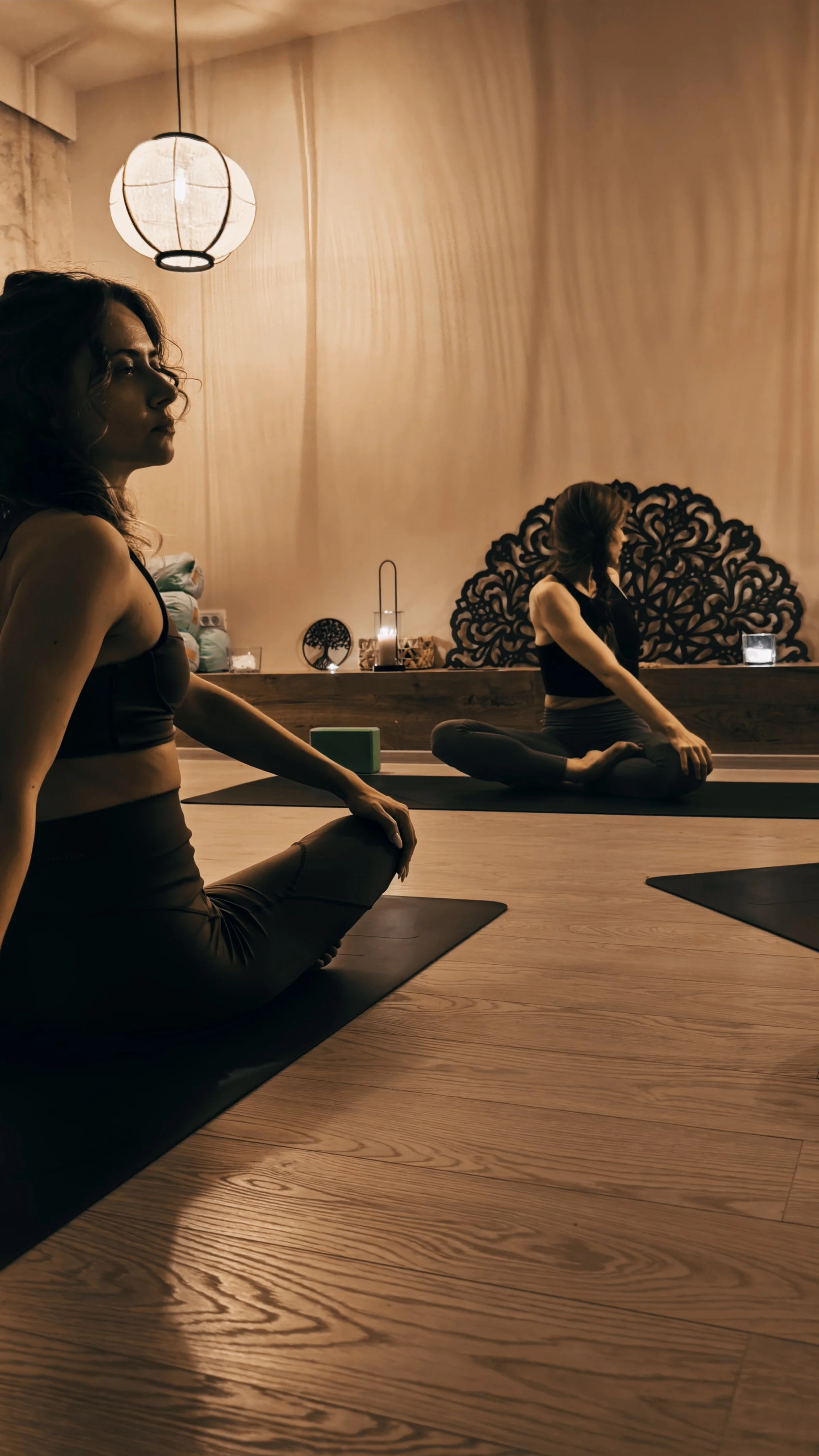 Two women practicing yoga in a dimly lit studio, sitting cross-legged on black mats with a decorative wooden wall piece in the background.