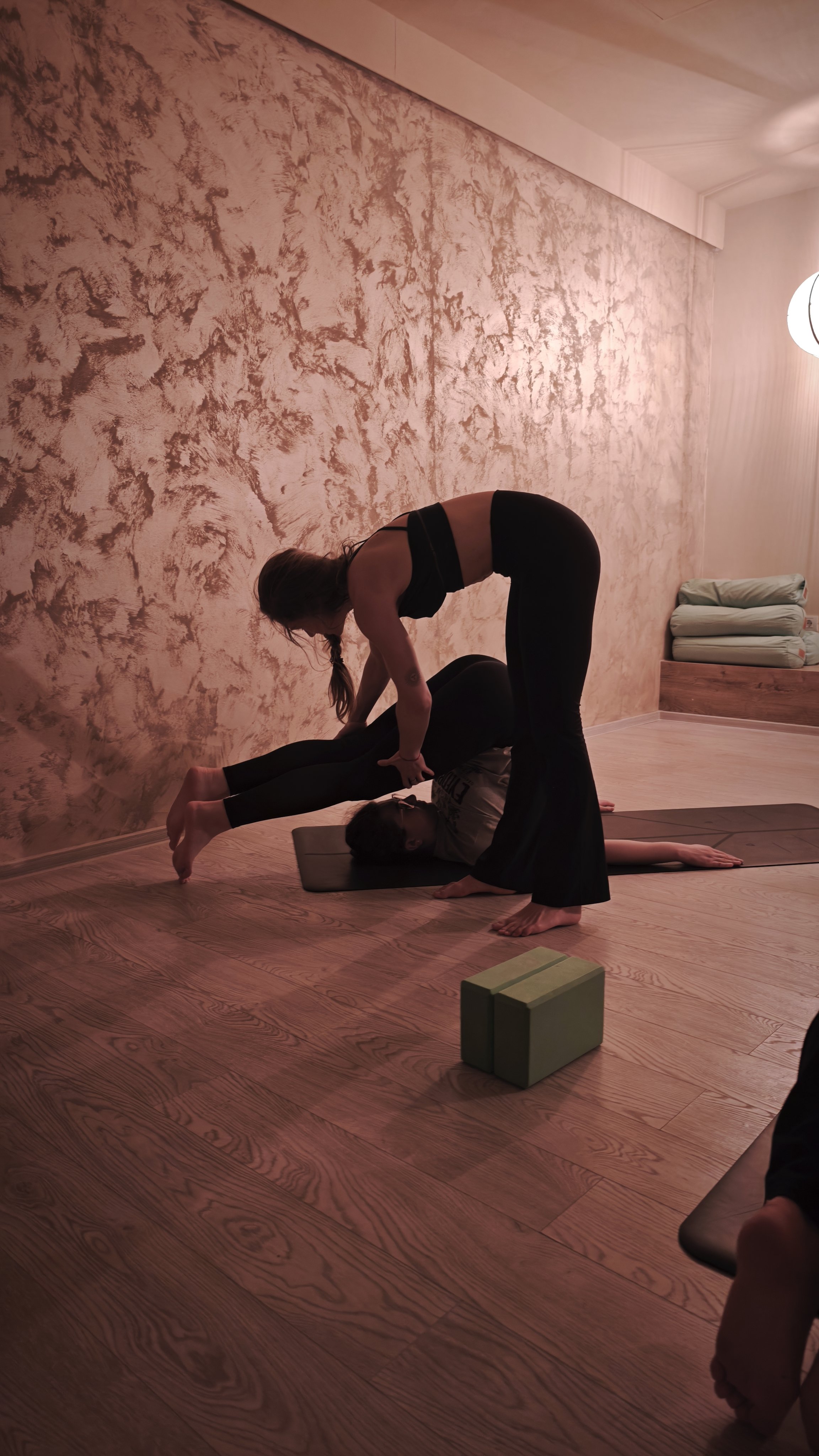 Two women practicing yoga indoors. One woman is lying on a yoga mat with another woman assisting her by positioning her ankles. The setting has warm lighting, textured wall, and wooden flooring.