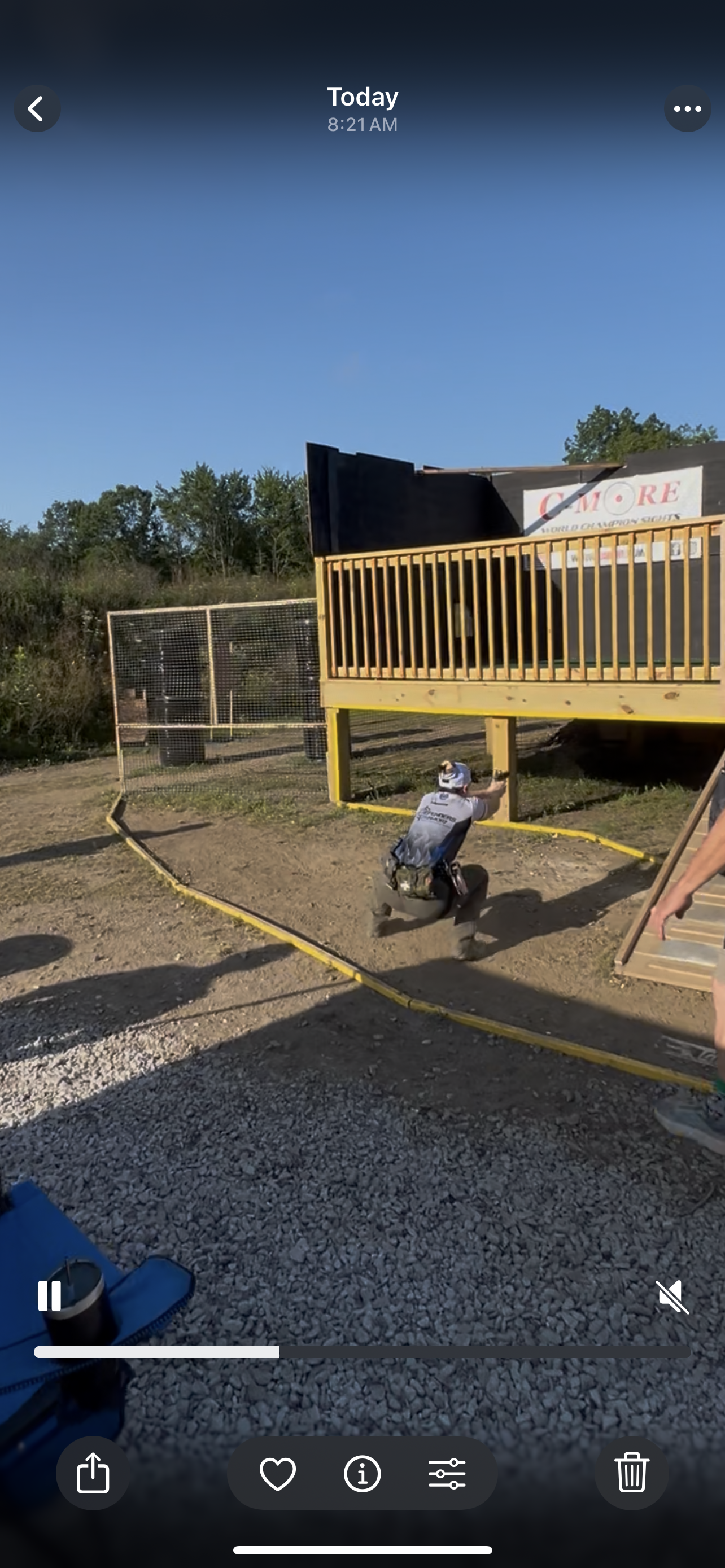 A person kneeling on the ground, aiming a firearm at a shooting range under an elevated wooden deck with a black backstop. The scene is outdoors on a sunny day with trees in the background.