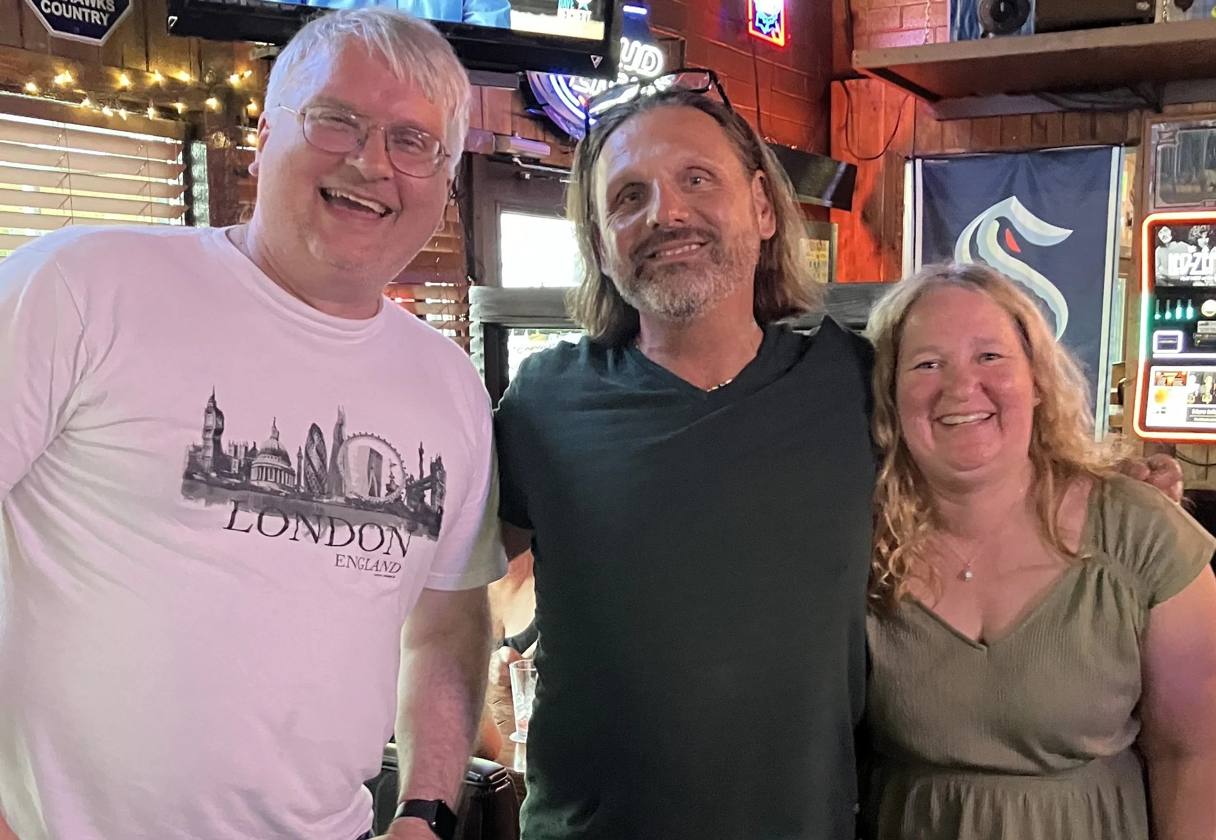 Three people standing close together in a bar or restaurant, smiling at the camera. The background has wooden decor, neon signs, and a window with blinds.