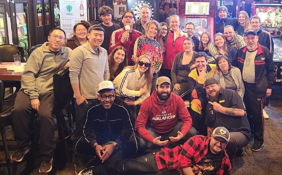 Group of smiling people gathered indoors, some sitting and some standing, posing for a group photo, with a woman holding a microphone in the center.