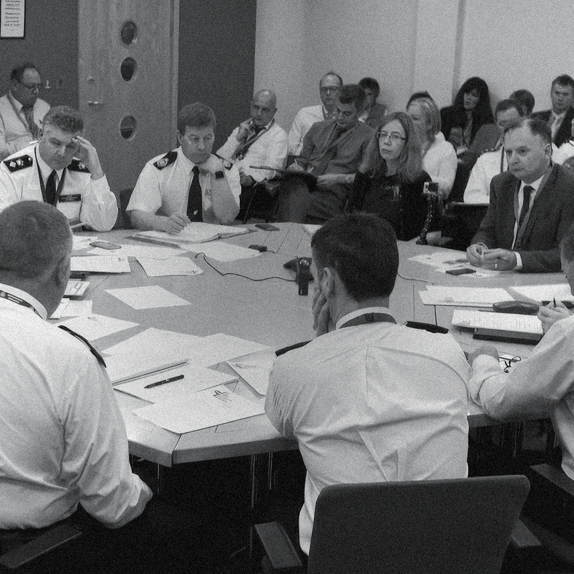 A black and white photo of a group of people in a serious meeting, some in uniform and others in business attire, seated around a table with documents.
