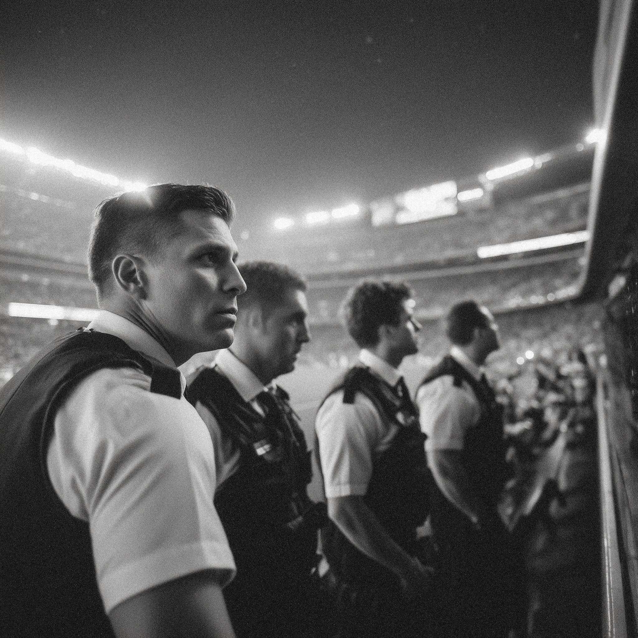 A black and white photo of four young men in uniform sitting in a control room or stadium, looking intently at monitors or screens, with a large stadium in the background.