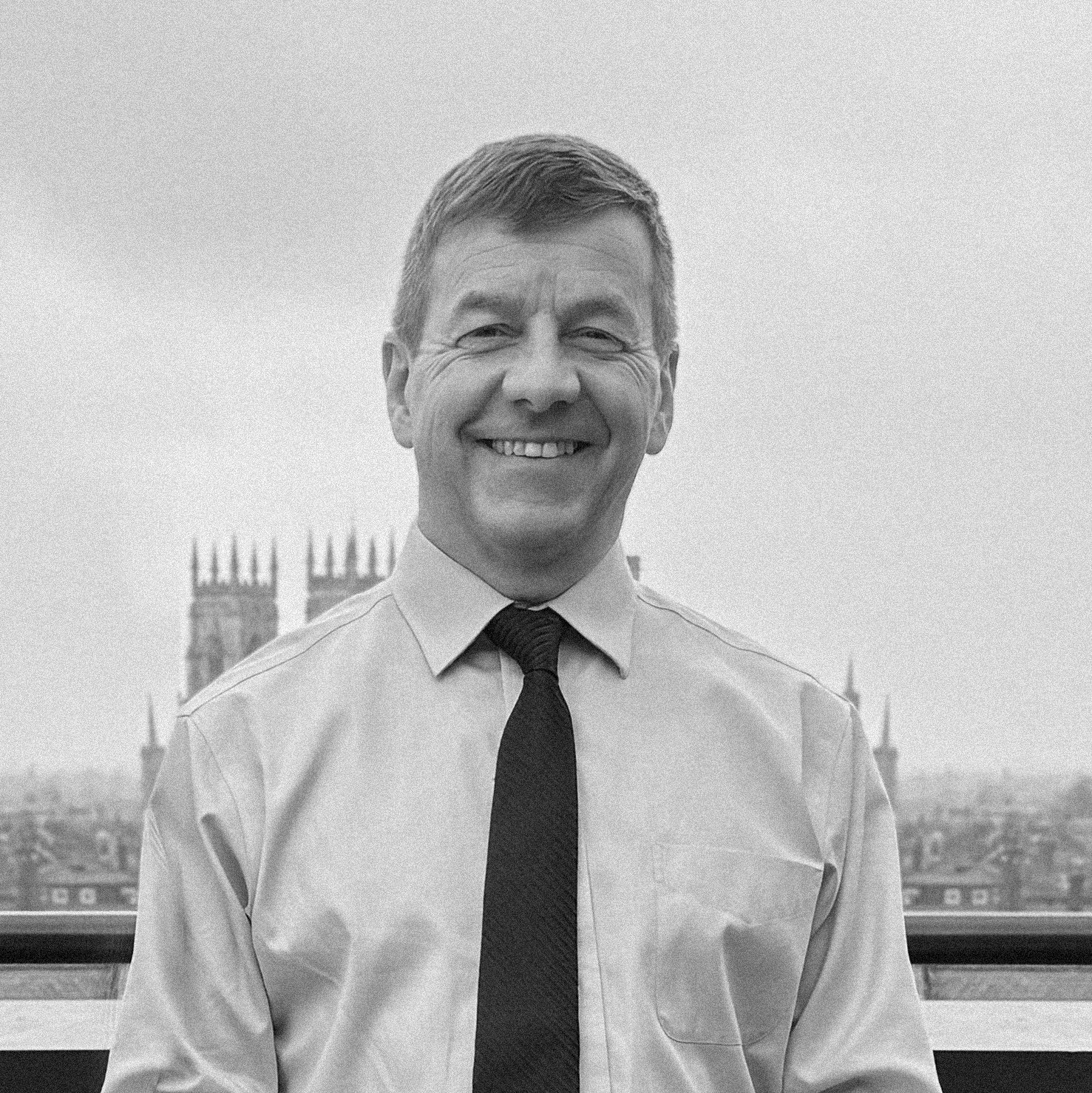 A smiling man in a dress shirt and tie standing outdoors with a cityscape and cathedral in the background.