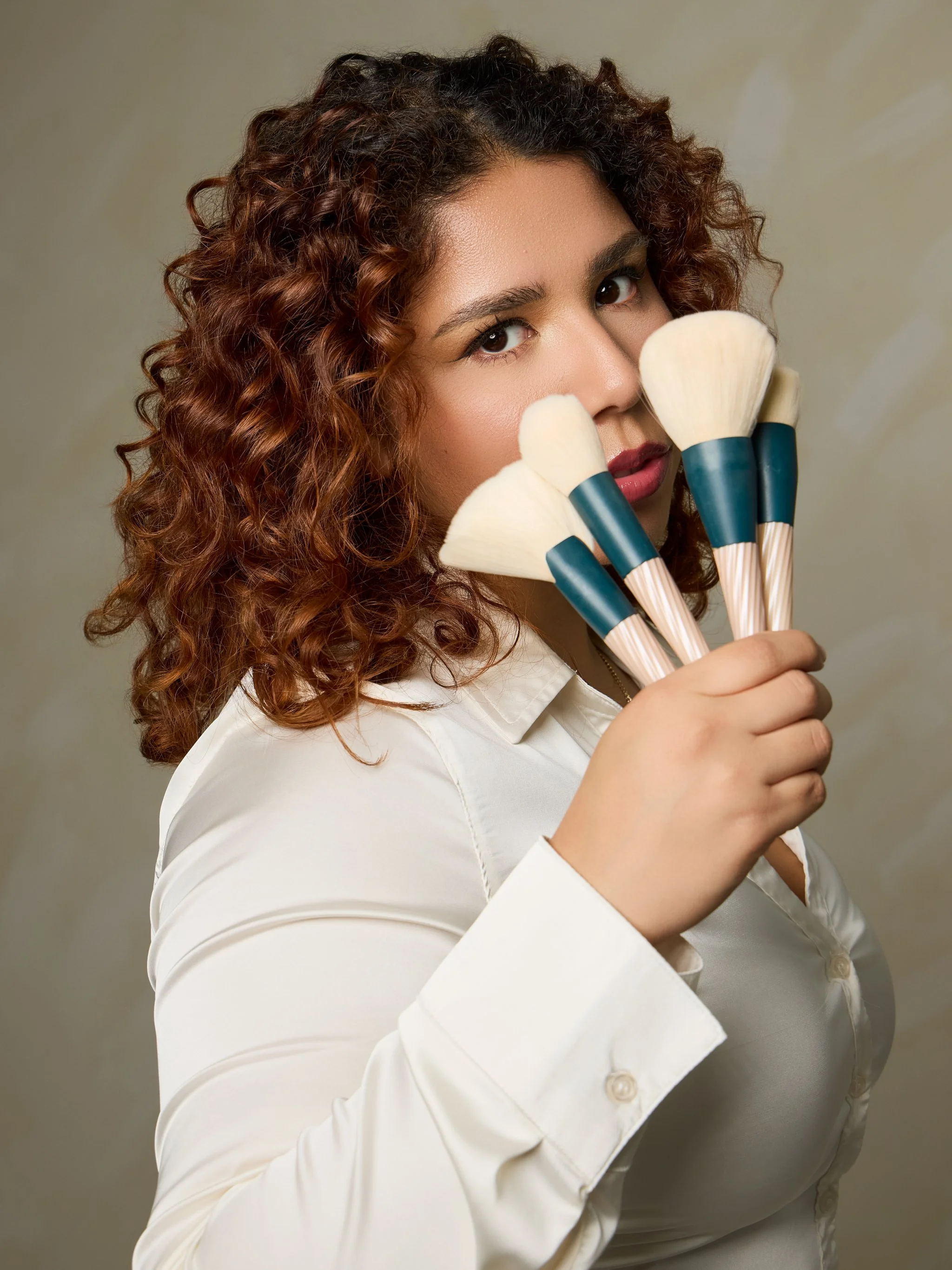A woman with curly brown hair holding four makeup brushes near her face, wearing a white shirt, against a light beige background. 
Castrian Makeup Studio; Emily Gonzalez; Atlanta, Georgia; Bridal Make-Up; Norcross, Spanish Make-Up Artist;