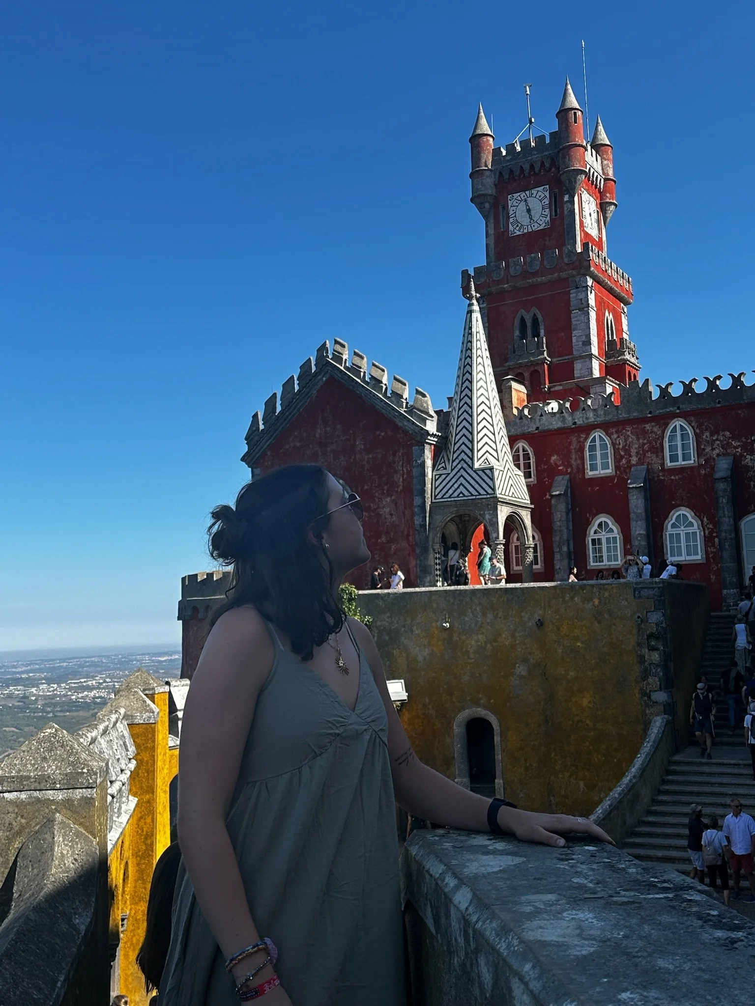 A woman standing on a stone balcony in front of a red castle with a clock tower, on a bright sunny day with clear blue sky.