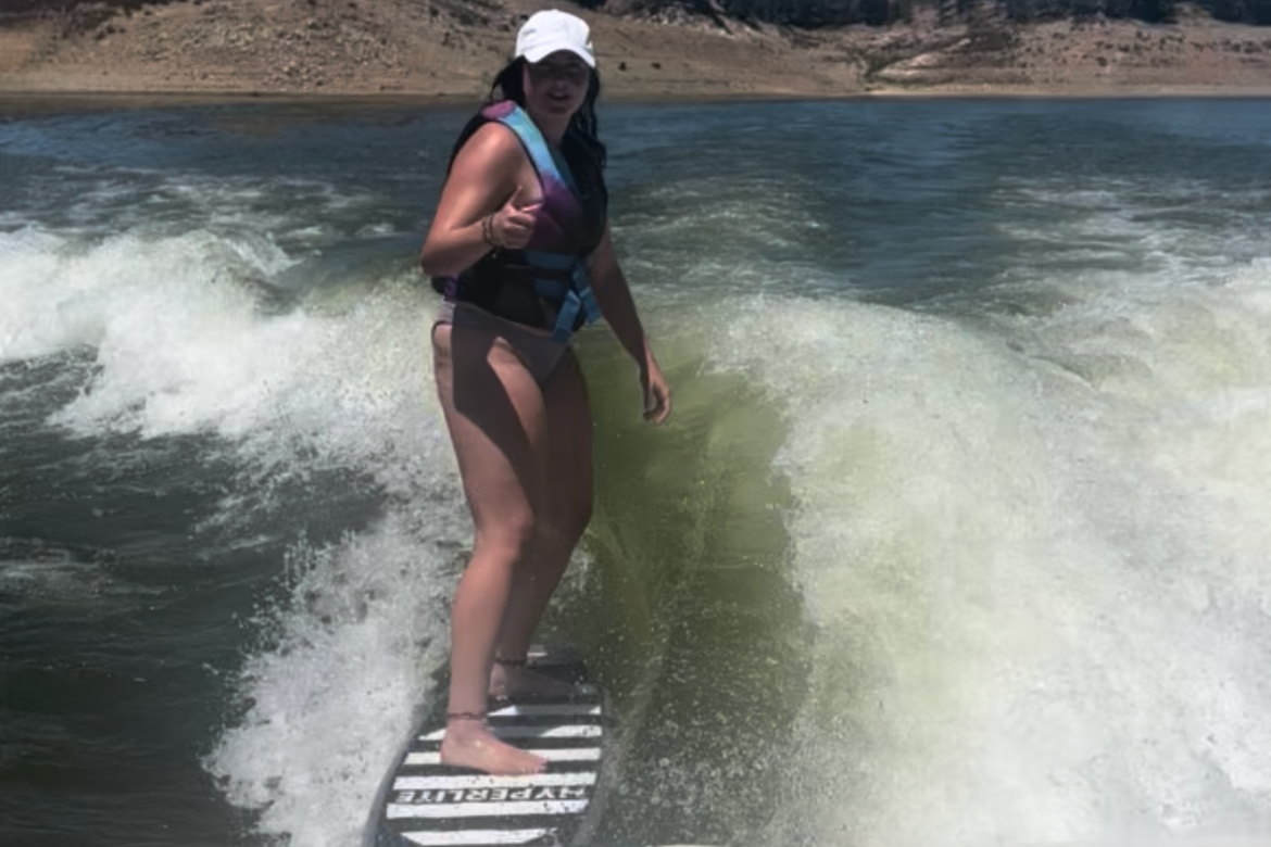 A woman surfing on a wave in a body of water, wearing a life jacket, hat, and swimsuit.