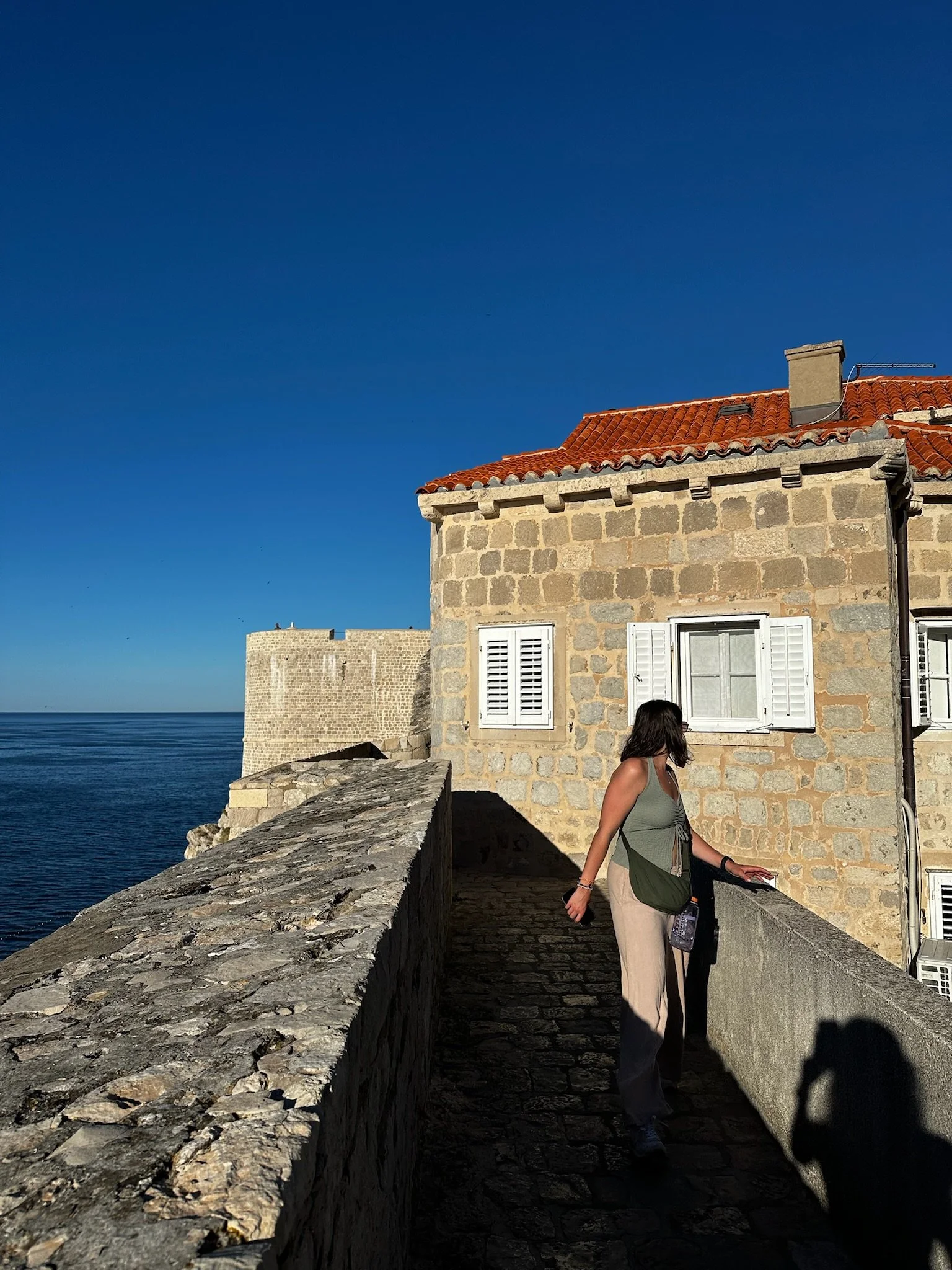 A woman with dark hair wearing a green top and beige pants walking along a stone pathway beside a stone building with white shutters, overlooking the sea under a clear blue sky.