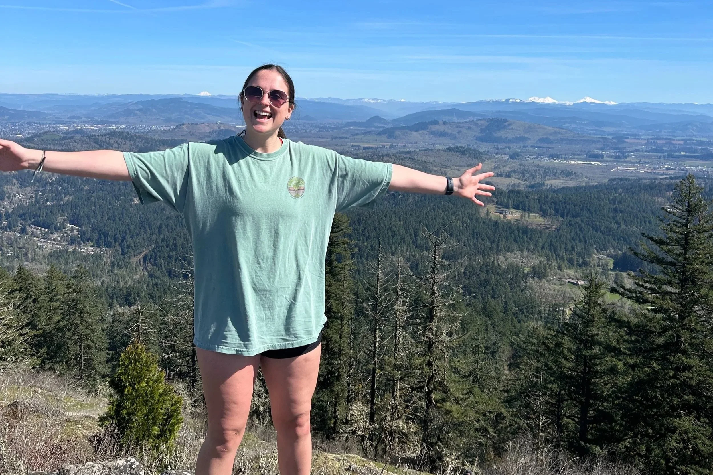 Woman standing on mountain with outstretched arms, wearing sunglasses, a light green T-shirt, and shorts, with a scenic view of forests, mountains, and a clear blue sky in the background.