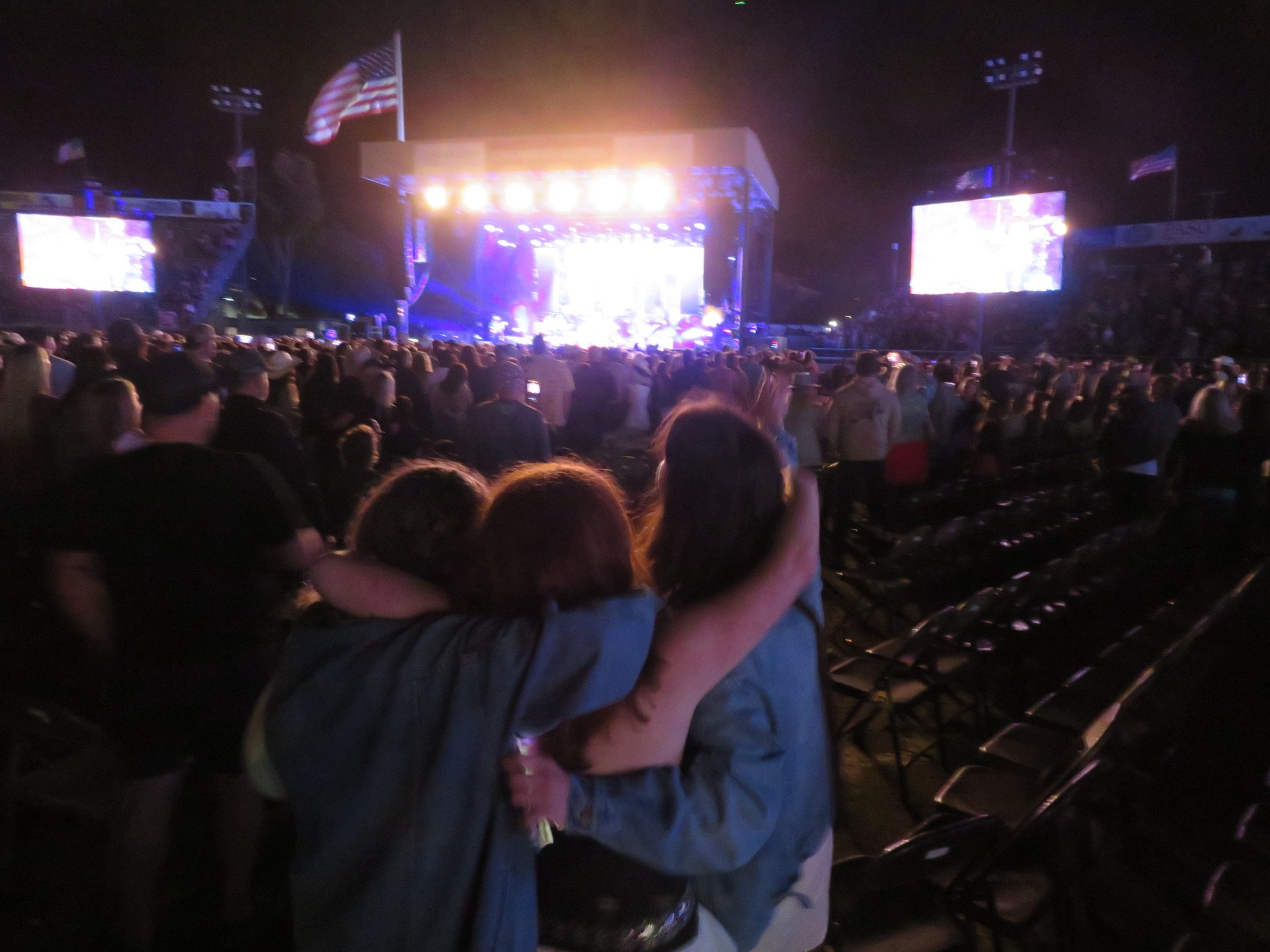 People hugging at a nighttime outdoor concert with bright stage lights, large screens, American flags, and a crowd in front of a stage.