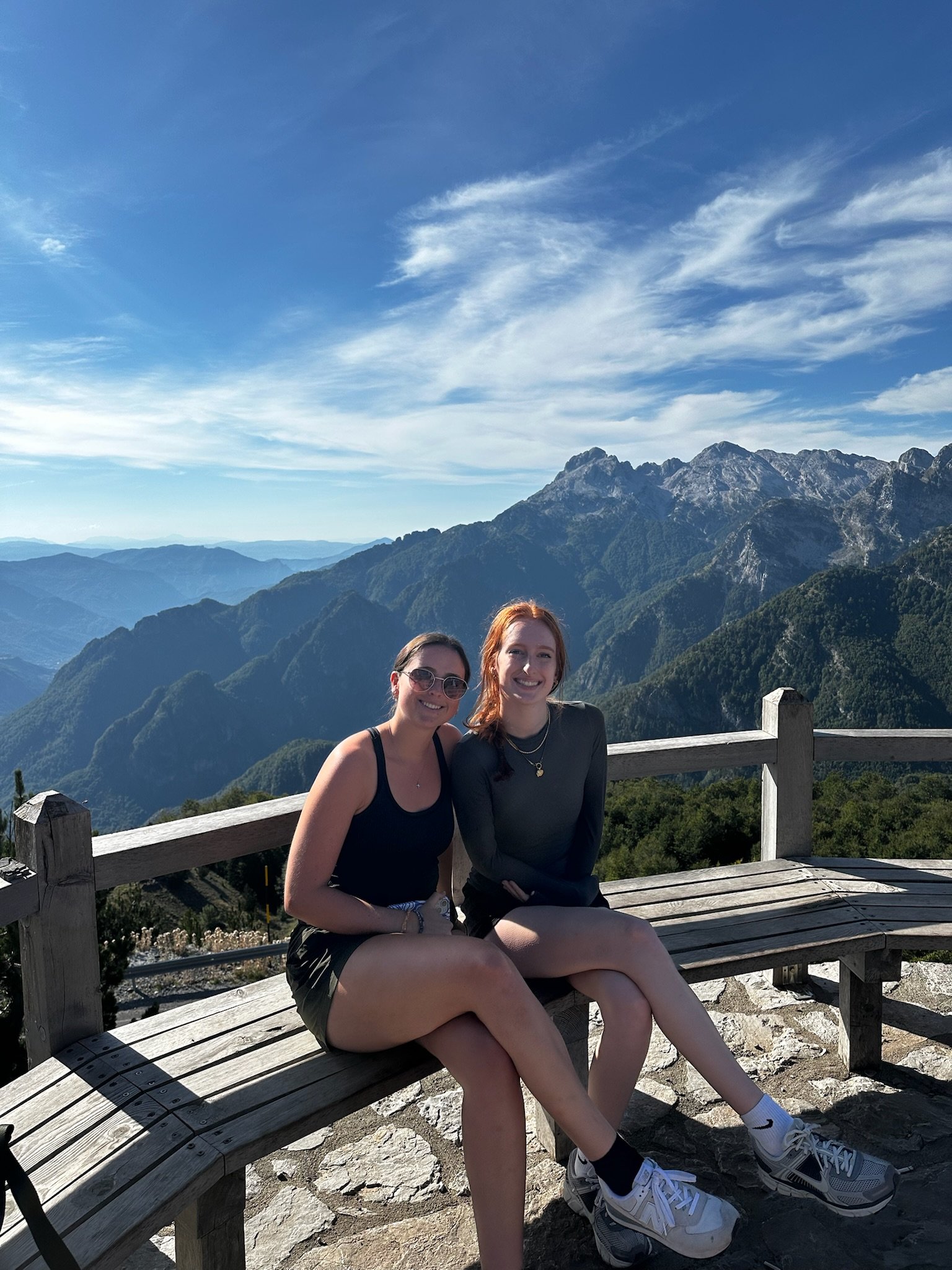 Two young women sitting on a wooden bench outdoors, smiling, with mountain scenery and a blue sky in the background.