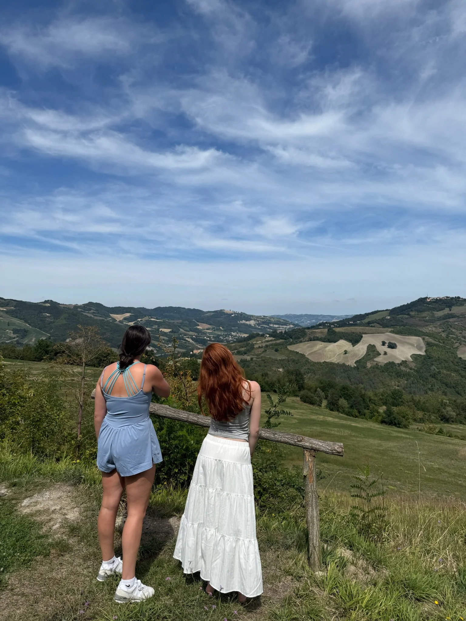 Two women stand near a wooden railing, looking out over rolling hills and valleys under a blue sky with wispy clouds.