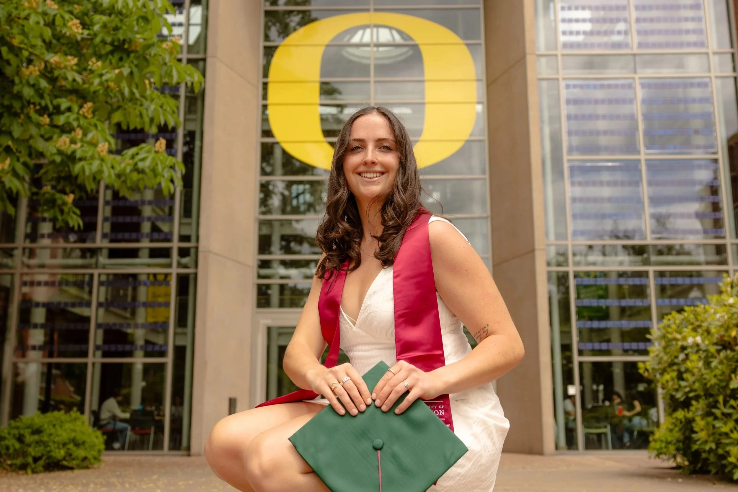 A young woman in a white dress with a red graduation sash, sitting outdoors in front of a modern glass building with a large yellow 'O' logo. She is holding a green graduation cap, smiling, with trees and bushes around her.