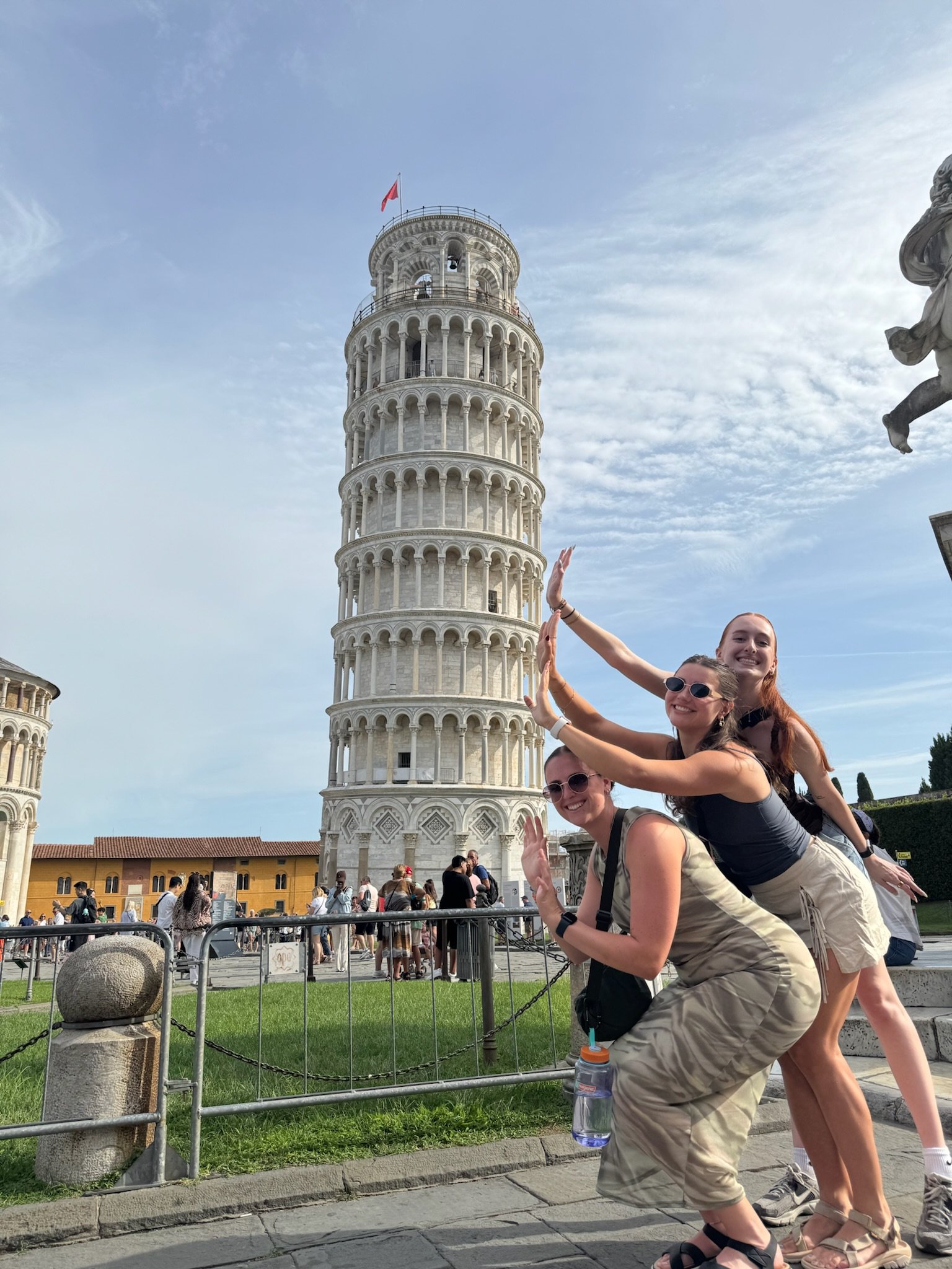 Three women posing in front of the Leaning Tower of Pisa in Italy, high-fiving each other.
